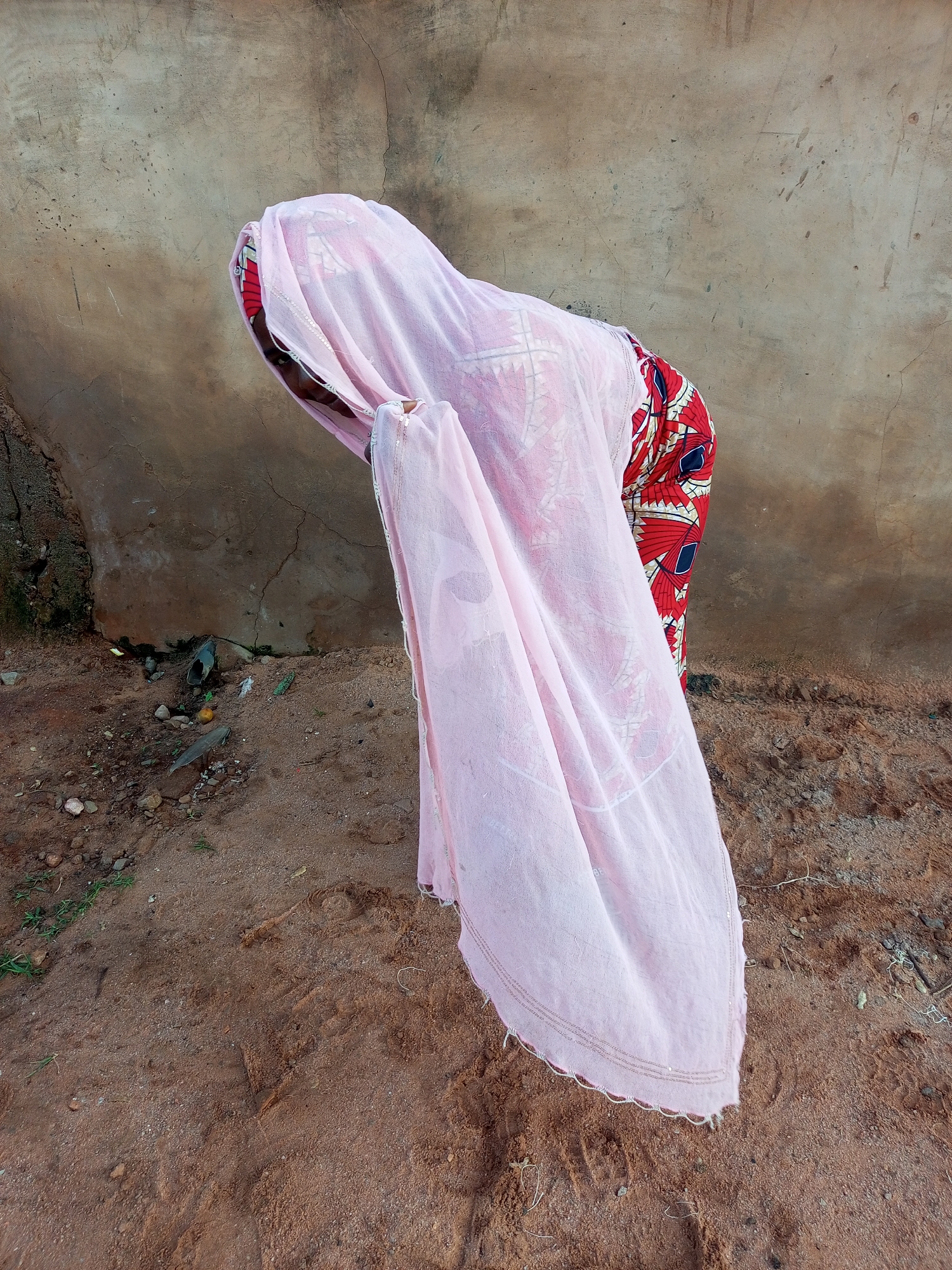 A person is shown from the side, bent over. They are wearing a pink veil that covers their head and upper body, and a patterned red, blue, and white dress or skirt. The person is standing on sandy ground in front of a textured, light-colored wall. The ground appears somewhat uneven and is covered in small stones and dirt. Some debris, including a plastic bag and what appears to be a small fruit, is visible on the ground near the wall. The scene suggests an outdoor environment, possibly a courtyard or an area adjacent to a building in a rural or semi-urban setting. No specific location within Tempure, Nigeria, can be identified from the visual information.