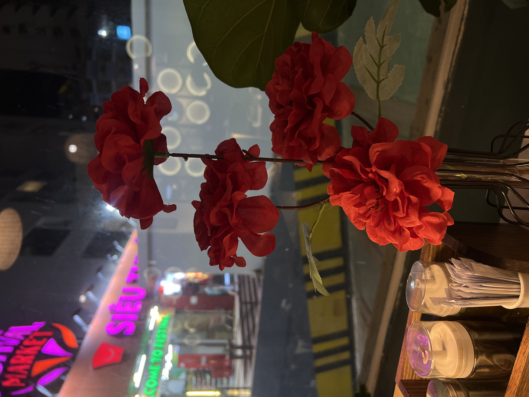 The image depicts a floral arrangement with red blossoms and green leaves in the foreground. Through the window, reflections of a Vietnamese street scene are visible. The signage "Siêu Thị Festival Market" is clearly seen in neon letters. The location is possibly Bắc Nha Trang, Vietnam, given the Vietnamese text. The background includes blurred lights and urban elements, implying a bustling commercial district. In the immediate foreground, there is a wooden rack holding cylindrical containers and paper products, suggesting this is a display in a store or restaurant.