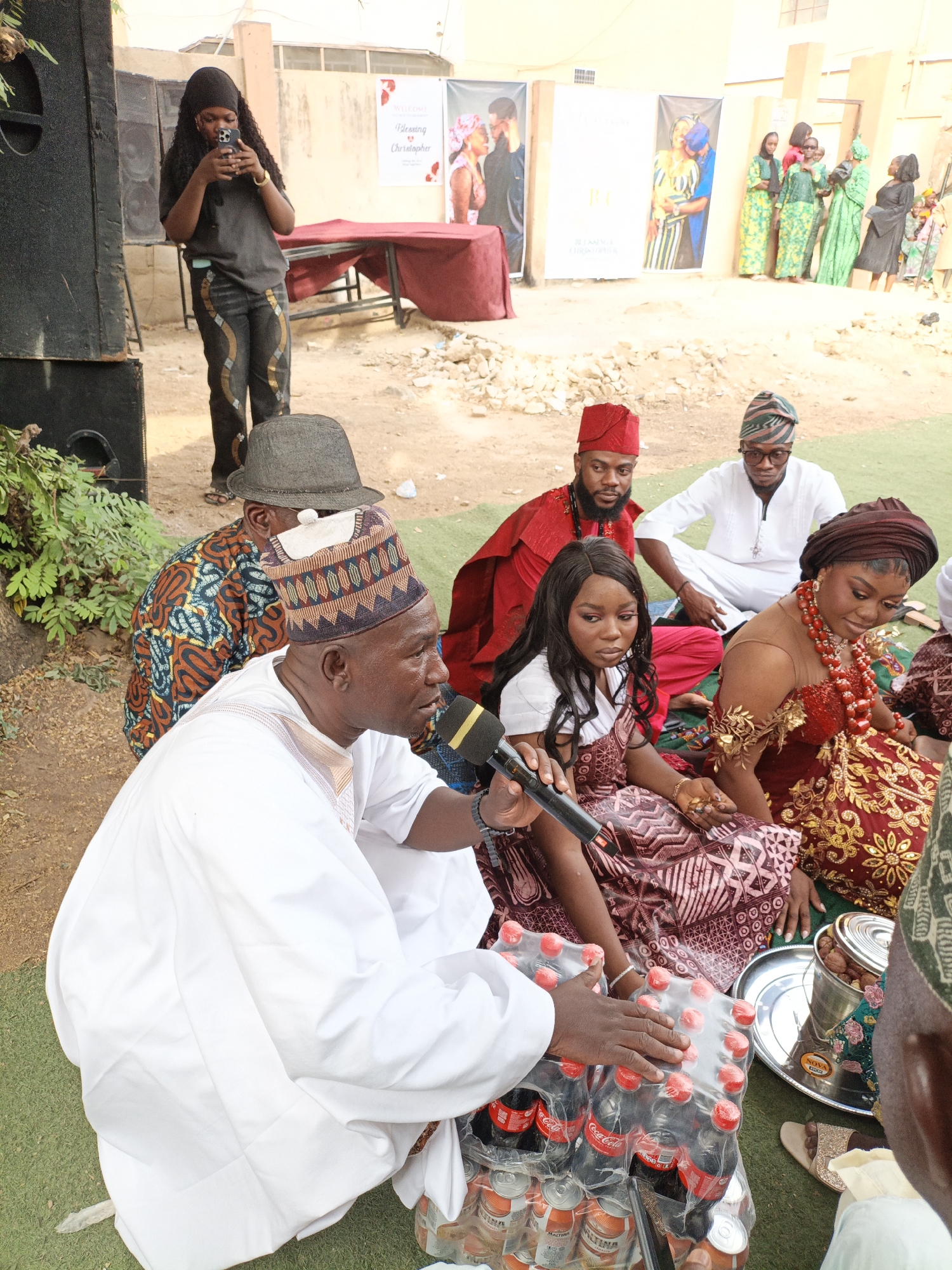 This image captures a vibrant traditional gathering, likely a marriage or engagement ceremony, in Sabon Gari West, Nigeria, set outdoors during daylight hours.
In the foreground, a man dressed in a white agbada and a patterned cap (fìlà) is seated on green artificial turf, speaking into a microphone. Beside him are multiple packs of Coca-Cola and Maltina soft drinks, as well as a silver tray holding nuts, possibly kola nuts, often used in traditional ceremonies. Seated close to him are two women in traditional attire: one in a dark patterned dress and another in an elaborate red and gold outfit adorned with red beaded jewelry. Both appear attentive, with the woman in red looking towards the speaker. Behind them, two men are seated, also in traditional Nigerian clothing, one in red attire and a red cap, and the other in white attire with a patterned cap.
In the mid-ground, a person with long dark hair, wearing a dark top and patterned trousers, stands near large black audio speakers, engrossed in their phone. The background features light-colored walls and several promotional banners. One banner prominently displays "WELCOME" and "Blessing Christopher," indicating the names associated with the event. Further in the background, a group of women in various colorful traditional dresses (green, yellow, and black) are standing against the wall, observing the proceedings. The ground transitions from the green turf where the main group is seated to bare, sandy earth with some rubble. The bright natural lighting suggests a clear, sunny day. The clothing and elements like the drinks and nuts strongly convey the cultural context of a Nigerian celebration.