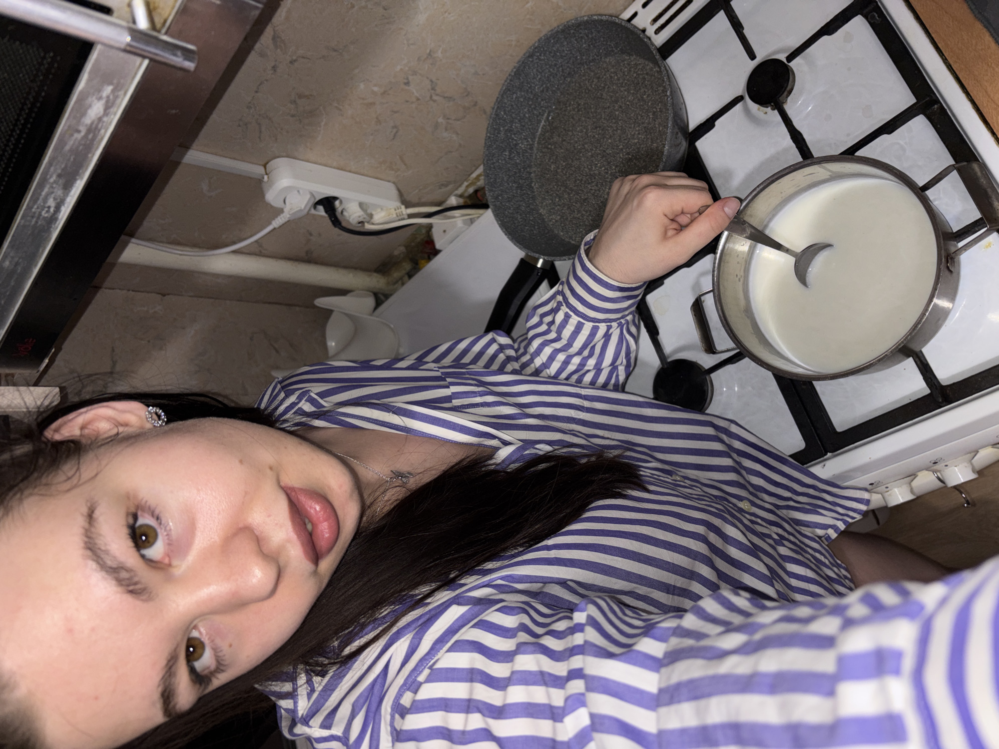 A woman is stirring milk in a pot on a gas stove in a kitchen. She is wearing a blue and white striped shirt. A frying pan sits nearby on the stove. The kitchen is located in Perm, Russia.