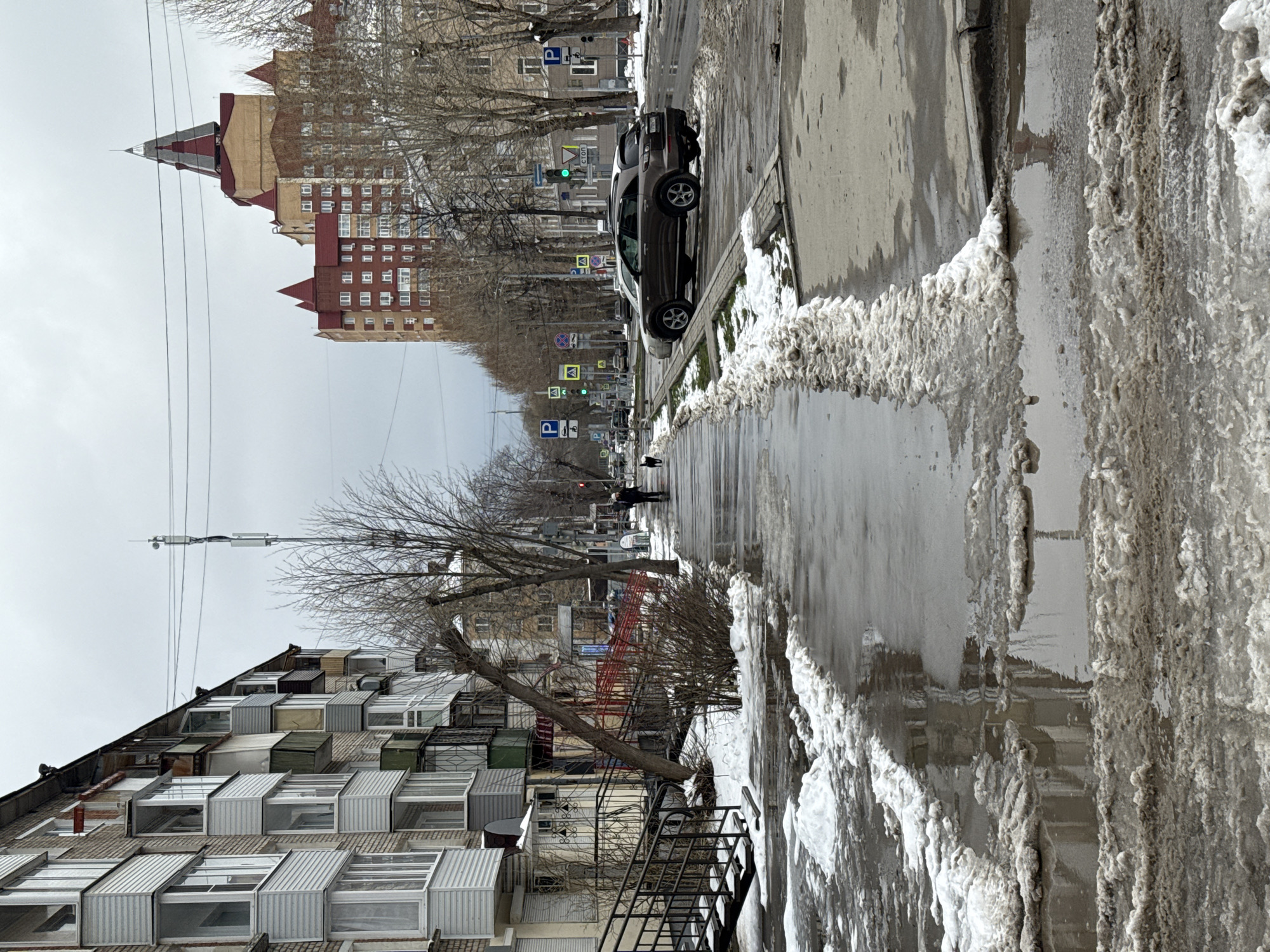 The image depicts a street scene in Perm, Russia, during a transitional weather period, likely late winter or early spring. The foreground features a partially snow-covered sidewalk with melting snow and slush along the curb. A multi-story apartment building is on the left, characterized by various enclosed balconies and a metal staircase leading up the side. On the right is the edge of a roadway with a parked dark-colored car facing away from the viewer. Beyond the car, the street extends into the distance, lined with leafless trees and buildings. A pedestrian is visible in the middle distance walking away from the viewer. Traffic signals are visible along the street. The sky is overcast and gray. A large apartment building with a uniquely-colored red roof is in the far background, giving away the location to be Perm, Russia.