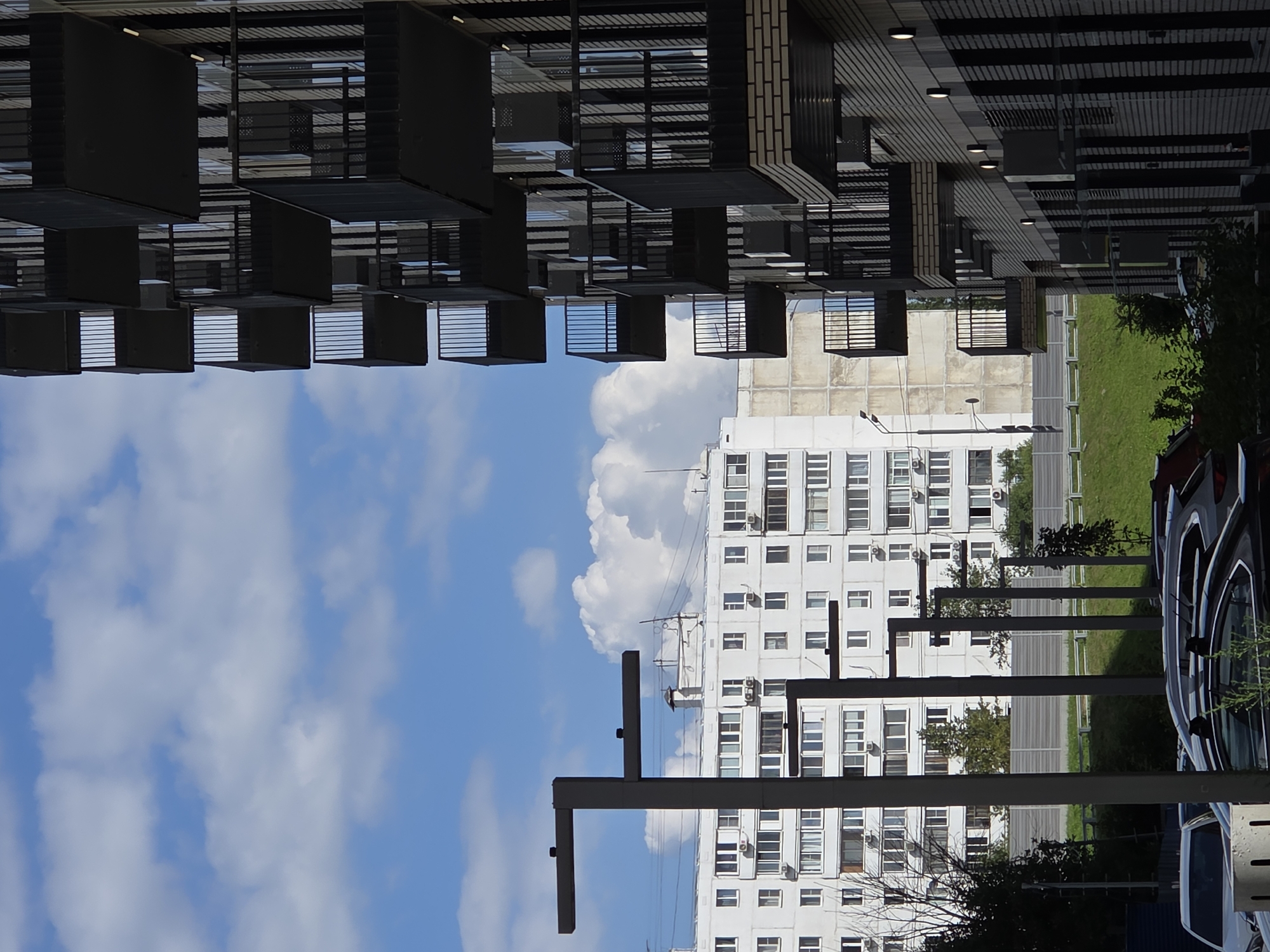 This is a photograph taken outdoors in Moscow, Russia, on a sunny day with a partly cloudy sky. The image features a modern apartment building with multiple balconies on the right side of the frame. These balconies are dark, likely black, and have metal railings. The building's facade appears to be made of light-colored brick with dark vertical accents.
To the left of the modern building, a section of an older, white apartment building is visible. This older building has numerous windows and what appear to be air conditioning units mounted on its exterior. In the foreground, several cars are parked, some of which are dark-colored. Streetlights or modern lamp posts are also present in the mid-ground. A grassy slope and a fence run behind the parked cars and extend towards the buildings. The sky is blue with fluffy white clouds. The overall scene depicts an urban residential area with a mix of older and newer architectural styles.