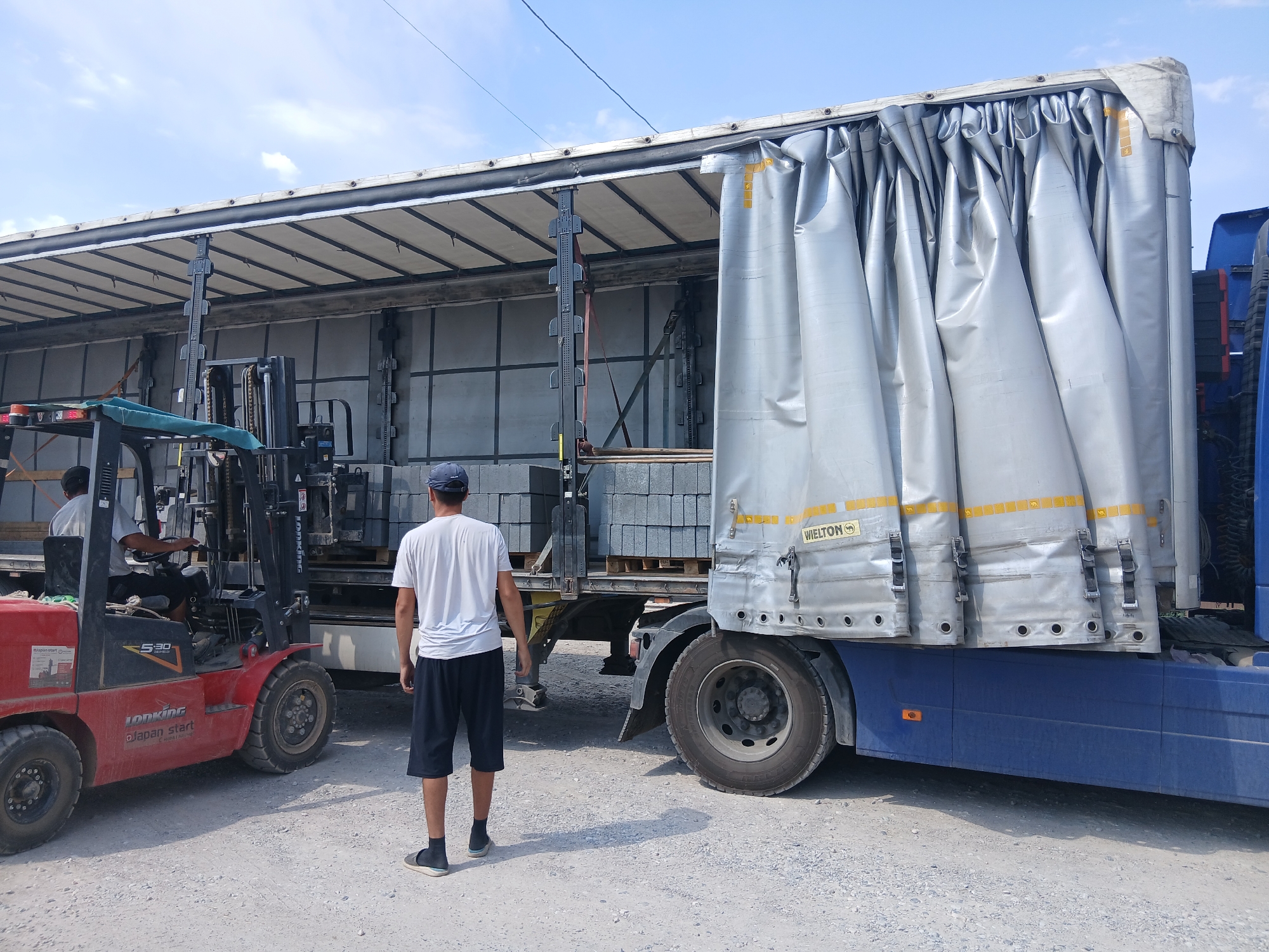 In Talas City, Kyrgyzstan, a man in a white t-shirt and black shorts stands facing away from the camera, observing a forklift operation. The forklift, a red "JONKING S30" model, is operated by another individual whose face is obscured by the vehicle's structure. The forklift is positioned near the rear of a large, silver-sided truck trailer, which is partially open. Pallets of gray, rectangular blocks, possibly construction materials, are visible within the trailer, suggesting a loading or unloading activity. The ground is covered in gravel and dirt, and the sky above is clear with a few scattered clouds. The scene appears to be an outdoor industrial or logistics area.