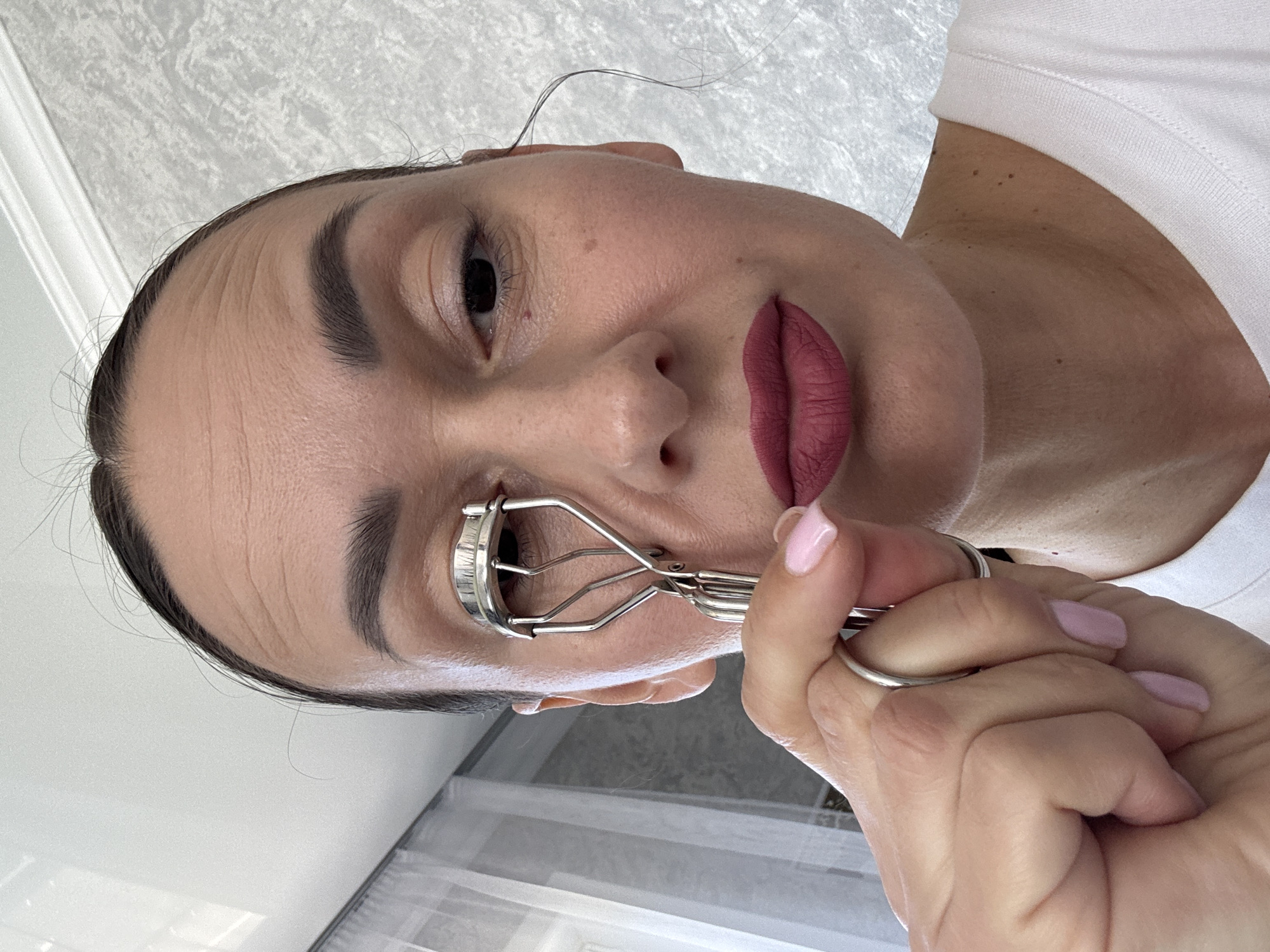 This close-up portrait, rotated 90 degrees counter-clockwise, features a woman in an indoor setting, likely in Sochi, Russia. She is shown from a low-angle perspective, looking upwards, possibly at the camera or a mirror.
**Subjects:** The central subject is a woman with dark hair pulled back, though a few wisps escape. Her face is well-lit, highlighting her features. She has dark, neatly shaped eyebrows. Her lips are adorned with a matte, dark pink or mauve lipstick. Her fingernails are painted a light pink. She is wearing a simple white t-shirt, visible on her upper body. On her right ring finger, she wears a silver-toned ring.
**Activity:** The woman is engaged in a beauty routine, specifically using a silver-toned eyelash curler on her left eye. Her right hand holds the curler in position.
**Scene/Setting:** The background features a light-colored wall with a subtle textured pattern, possibly wallpaper. A white architectural element, perhaps a window or door frame, is also visible. The bright, even lighting suggests it is daytime.
**Notable Details:** The woman's skin displays natural texture, including faint lines on her forehead and several small moles or freckles on her cheek and neck/chest area. Her expression is neutral with a slight, subtle smile. The image has a personal, candid feel, focusing on the intimate act of self-care.
**Visible Text:** There is no visible text in the image.
