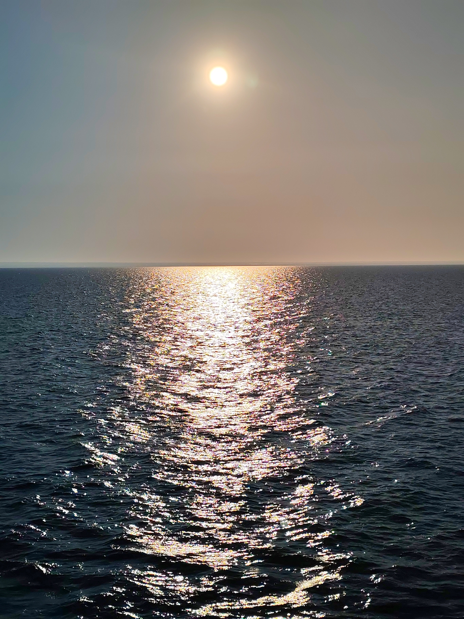 The image depicts a body of water under a bright sun. The sun is positioned in the upper center of the frame, casting a strong reflection on the water's surface, creating a shimmering path of light that stretches towards the viewer. The water appears dark and choppy, with small waves visible. The horizon line is clearly defined, separating the water from a hazy sky that transitions from a light grey to a pale blue. No people, animals, or man-made structures are visible in the image. The scene appears to be a natural landscape, likely an ocean, sea, or large lake. There is no indication of the city of Prislon', Russia, in the image.