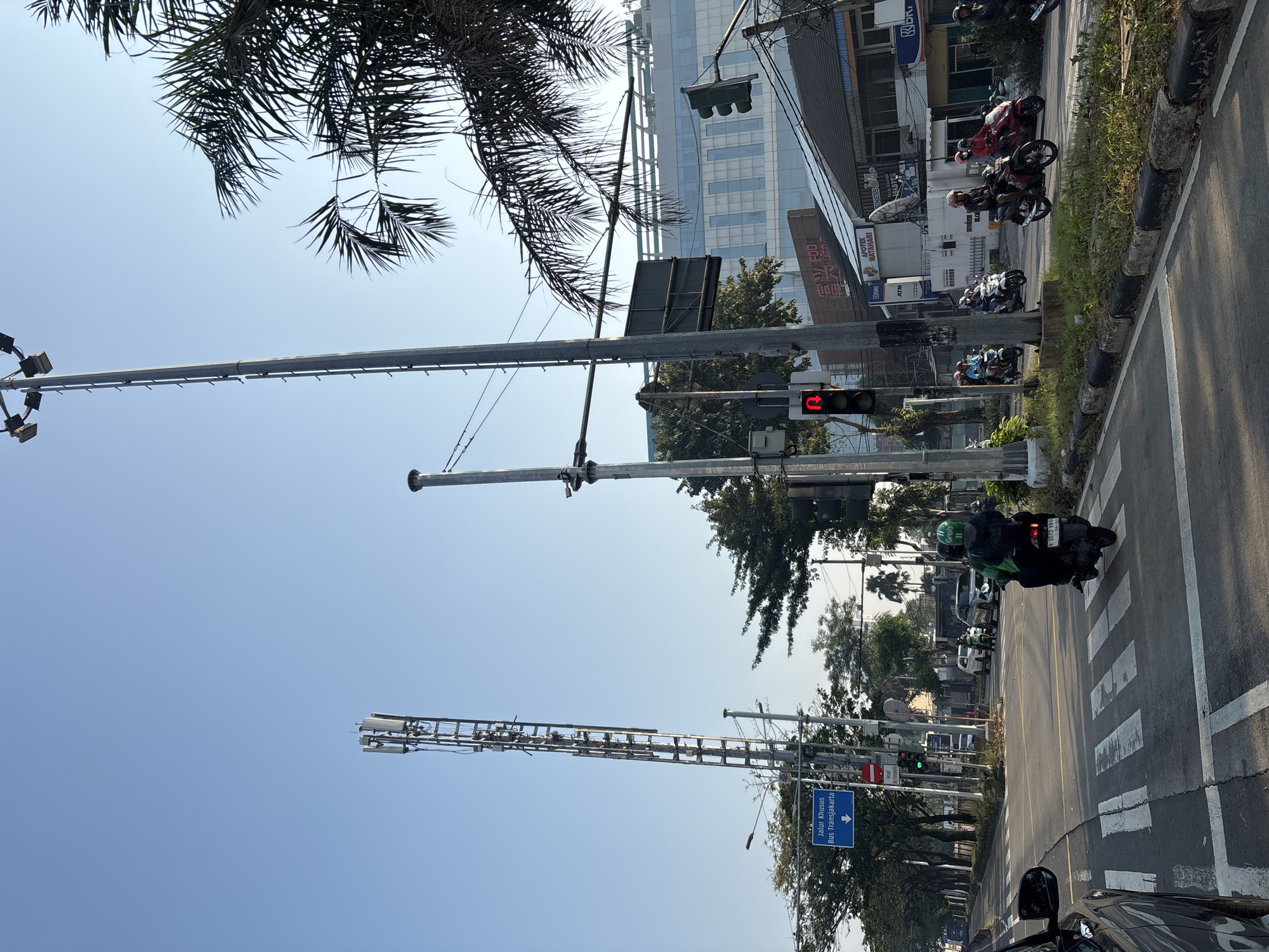 In West Jakarta, Indonesia, a street scene unfolds under a clear blue sky. A tall telecommunications tower stands to the left, its metal lattice structure supporting multiple antennas. A prominent blue sign with white text, "Jalur Khusus Bus Transjakarta," indicating a dedicated bus lane, is visible below the tower, accompanied by a downward arrow. A traffic light shows a red signal. Several motorcycles, some with riders wearing green helmets and jackets, are present on the road, with one on a pedestrian crossing. A large building with a modern glass facade rises in the background. Palm trees and other foliage line the sides of the road. The scene captures a typical urban traffic environment with active transportation.