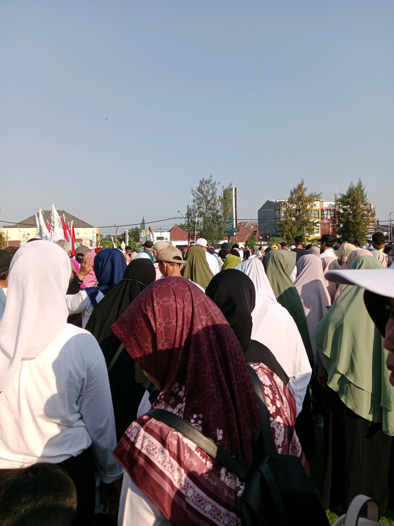 This is a medium shot, taken from a low angle, of a crowd of people standing outdoors. The sky is clear and blue, and the sun is shining. In the background, there are buildings and trees. The people in the foreground are mostly women, wearing headscarves and traditional clothing. They appear to be attending some kind of religious gathering or event. The image captures a sense of community and shared experience. The crowd is densely packed, suggesting a large turnout. The overall impression is one of a vibrant and active community.