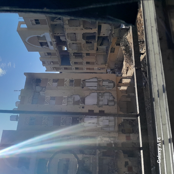 This image captures a scene of extensive urban destruction in Khan Yunis, Palestinian Territory. The foreground is partially obscured by a dark, horizontal bar, likely a window frame or railing, through which two multi-story residential or commercial buildings are visible under a clear blue sky with sparse white clouds. Sunlight emanates from the upper left, creating lens flare.
The primary building on the left is a light-colored, multi-story structure, at least six floors high, exhibiting severe damage. Its facade is pockmarked with numerous impact holes, and large sections of exterior walls are missing, exposing internal rooms and structural elements. Many windows are shattered or completely absent, though some retain decorative arched frames. Burn marks are visible on upper floors. Water tanks are present on the roof.
To the right and slightly behind, a second, similarly constructed light-colored multi-story building also shows significant structural damage. Large sections of its facade are collapsed or missing, revealing multiple floors of interior devastation. An arched architectural detail is visible on an upper floor, also damaged.
Between these two buildings, the ground level is covered with extensive piles of rubble, concrete debris, and scattered refuse, indicative of collapsed structures. A single individual, dressed in dark clothing, is observed walking on the debris-strewn ground between the buildings, near the center-right. No other discernible people, actions, or specific objects are clearly identifiable within the scene, which is dominated by the widespread structural damage to the buildings and the accumulation of debris.