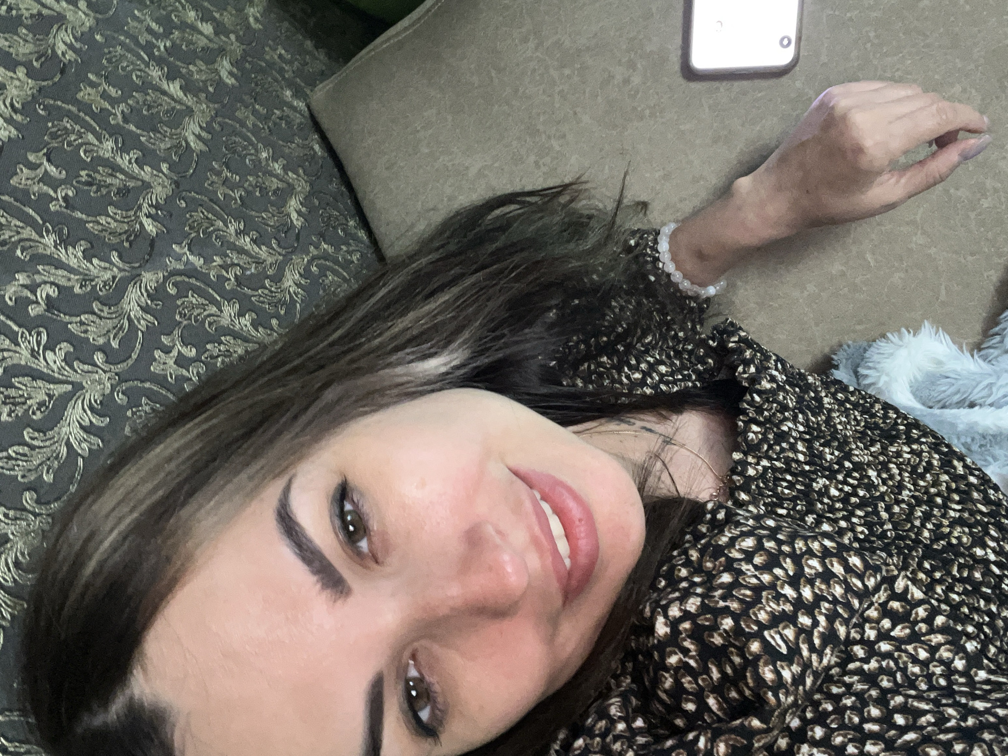 A close-up, overhead shot shows a woman with dark hair with lighter streaks, brown eyes, and full lips, smiling at the camera. She is wearing a black and brown animal print top with smocking across the chest and shoulders. A delicate necklace is visible around her neck. Her right arm is resting on a textured, tan surface, and she is wearing a white beaded bracelet. To the right of her arm, a smartphone with a bright screen is partially visible. The background consists of a dark, patterned wallpaper.