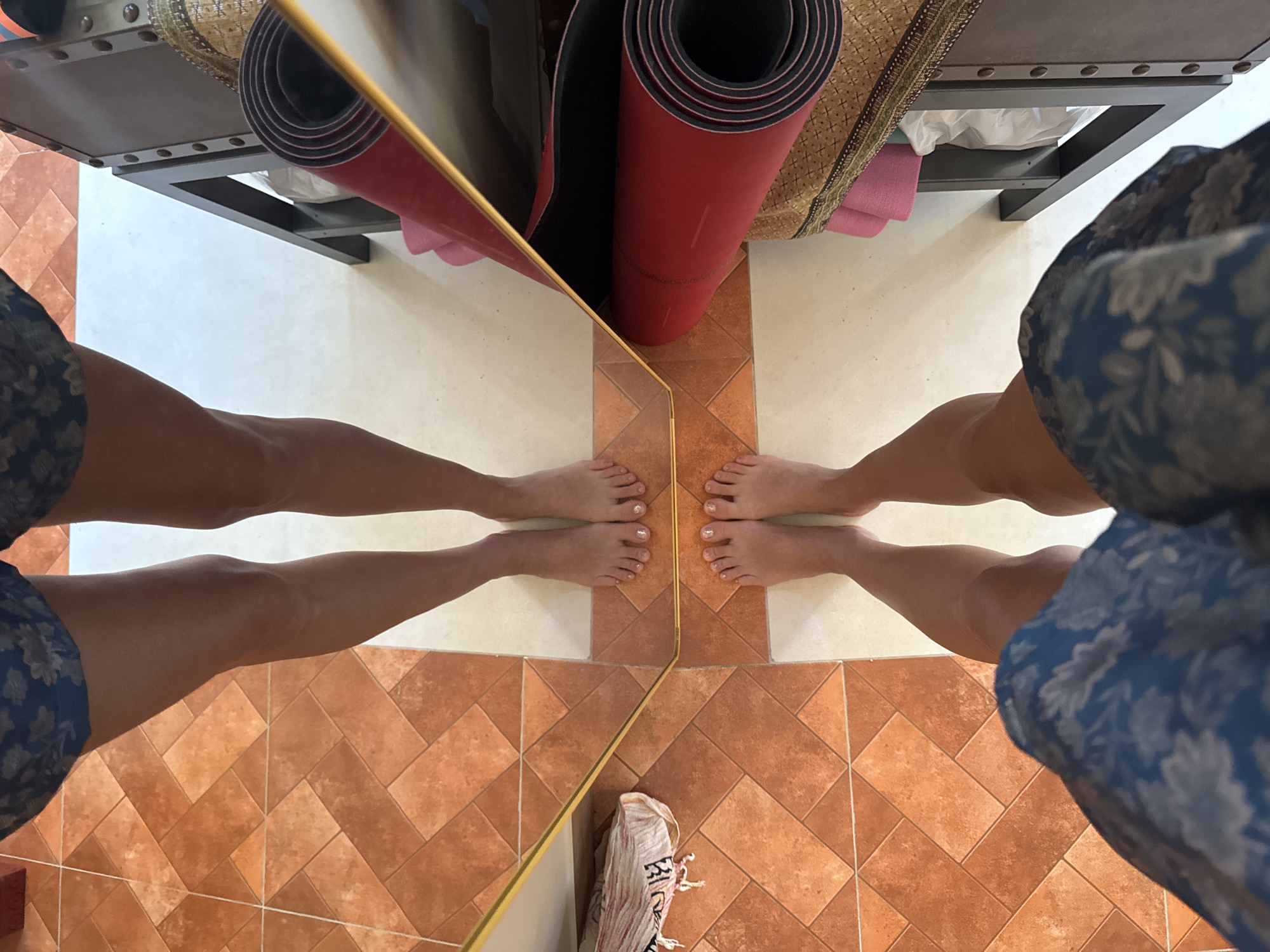 The image shows a first-person perspective of a person standing on a tiled floor with a mirror reflecting their legs and feet. The floor is a mixture of orange-brown and white tiles. To the right of the reflection, a dark metal shelf holds several rolled-up yoga mats, predominantly red and pink, along with some fabric. A white shoe with a pink sole is visible on the floor to the left of the reflection. The person is wearing blue shorts with a floral pattern. The scene appears to be indoors, likely in a room that might be used for yoga or exercise, possibly in a resort or hotel in Mũi Né, Vietnam, given the presence of yoga mats and the general ambiance. The lighting suggests it is daytime.