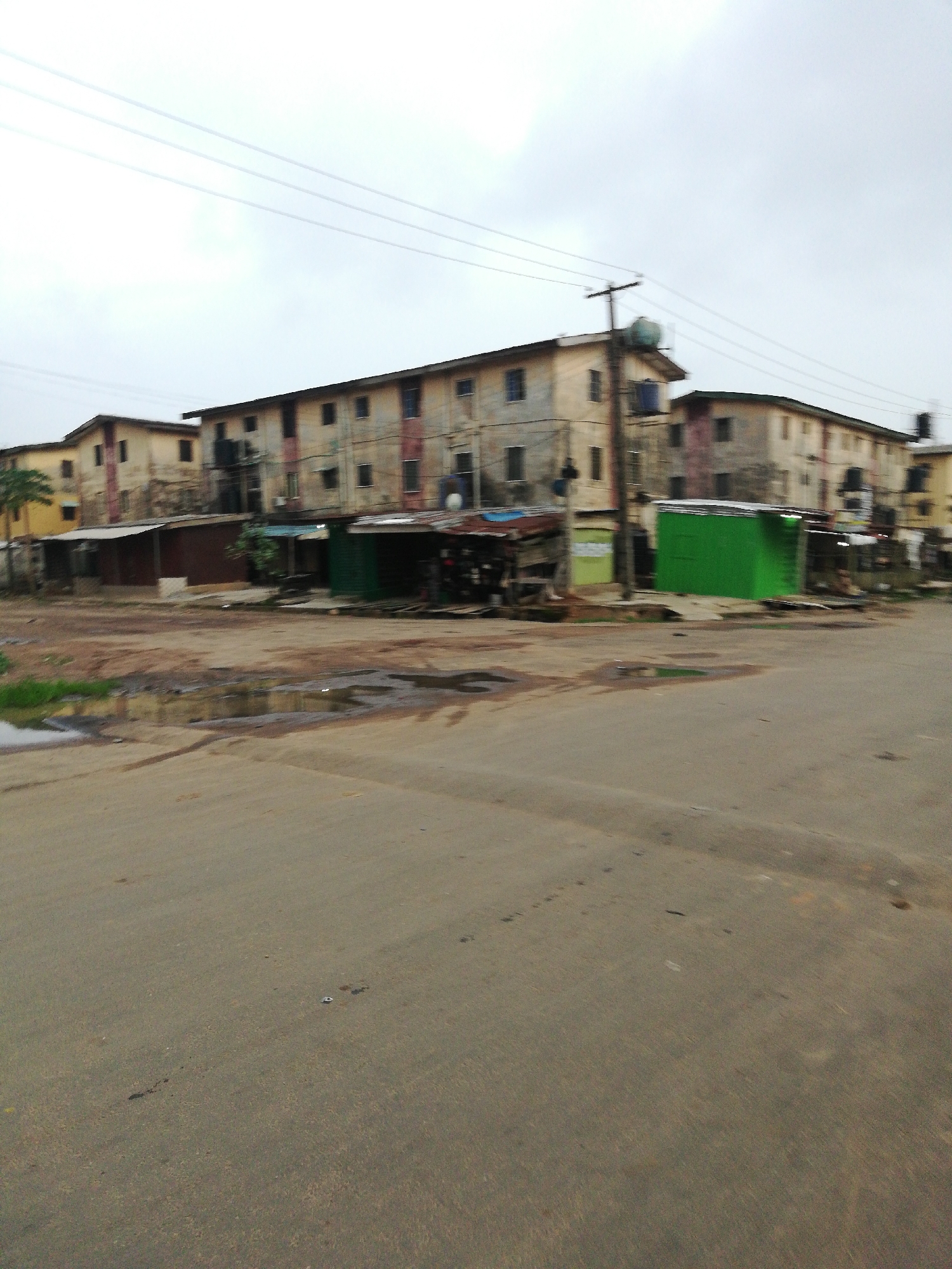 The image depicts a street scene at a road intersection in an urban area. The buildings, likely apartment blocks, show signs of disrepair with fading paint and visible stains. Along the base of the buildings, there are makeshift shops or stalls. One is painted green, another brown. Power lines are visible above. The road surface is paved but has some patches of standing water and dirt. The sky is overcast, suggesting a possibly humid or rainy climate. There are no people immediately visible.