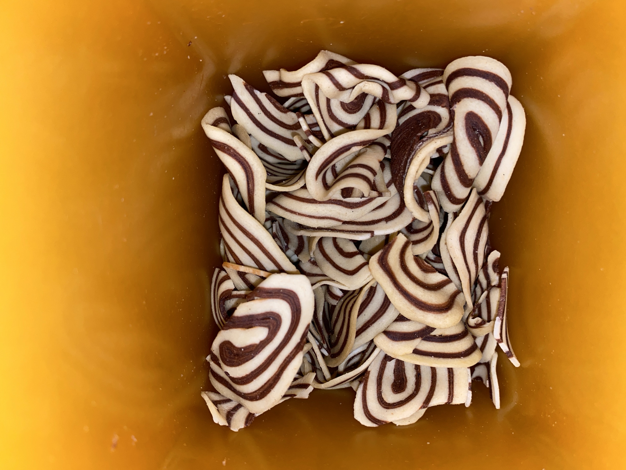 The image shows a top-down view of a container filled with striped chocolate and vanilla twisted cookies. The container appears to be made of yellow plastic. The cookies are arranged haphazardly within the container, exhibiting alternating dark and light brown stripes in a swirl pattern. No people, specific actions, or environmental location in Lhokseumawe, Indonesia, are discernible from the image.