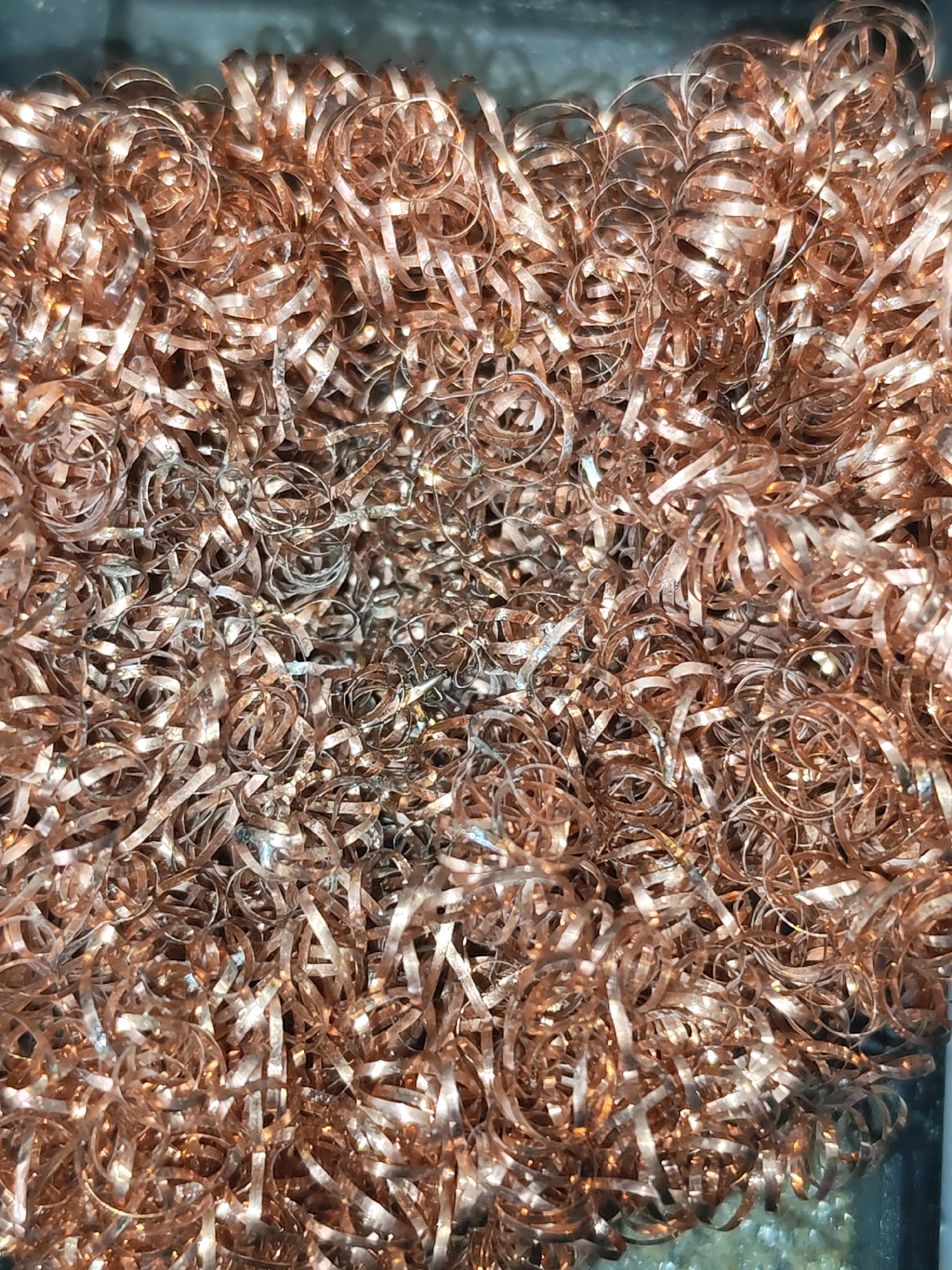 The image shows a close-up, overhead view of a pile of copper shavings. The shavings are thin, ribbon-like pieces of copper, many of which are curled into spirals or loops. They have a metallic sheen and range in color from bright copper to a duller, slightly oxidized brown. The texture appears to be rough and irregular due to the nature of the shavings. The lighting highlights the reflective surfaces of the metal, creating bright spots and shadows within the pile. There is no discernible background detail other than what appears to be a dark, possibly metallic or plastic edge on the left side of the frame, suggesting the shavings are contained within something. There are no people or discernible actions taking place in the image. The image is a still shot of static material. There is no information in the image that can be used to determine its location in the city of Izhevsk, Russia.