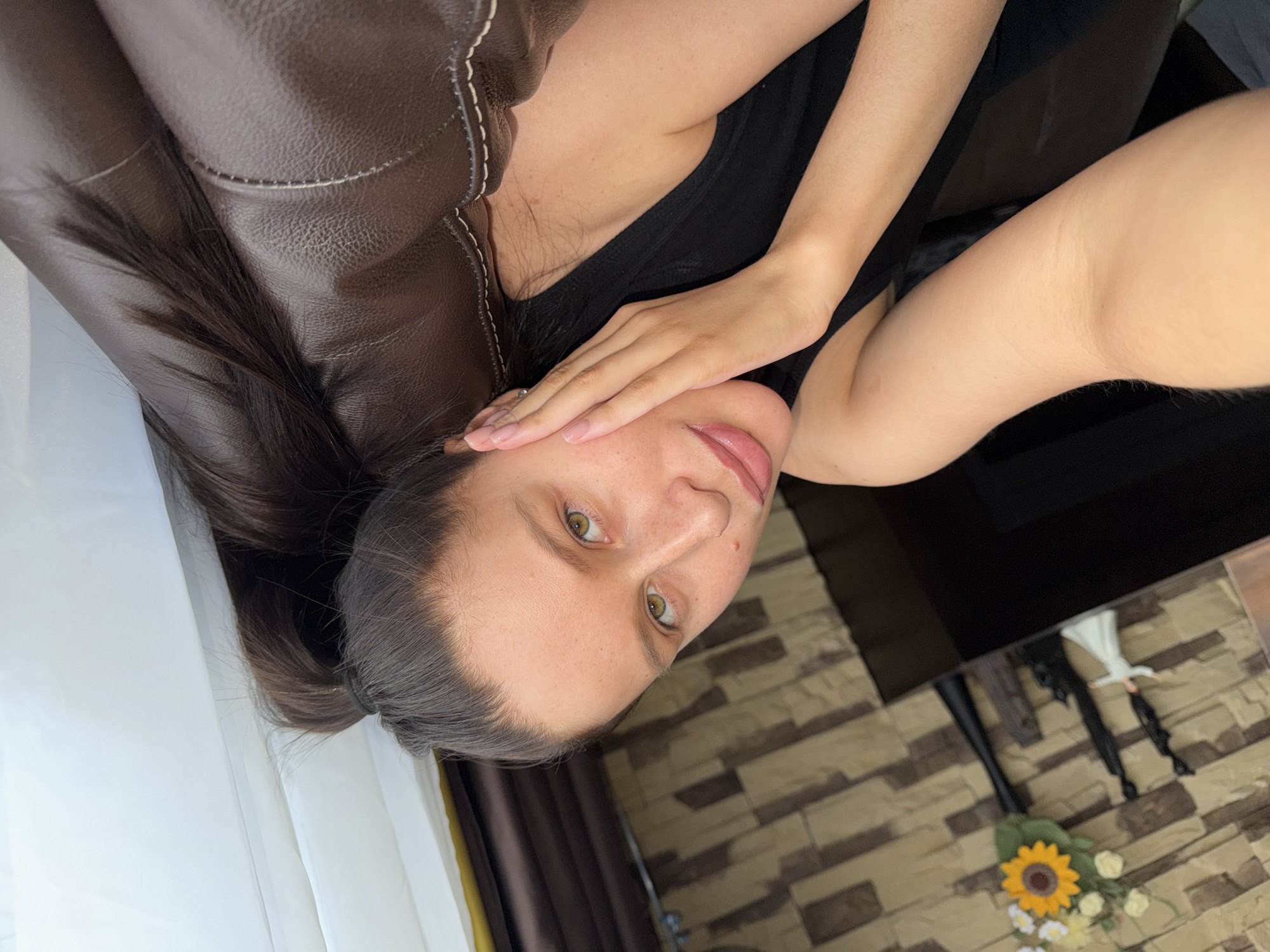 A young woman is depicted in a selfie, lying on her back indoors, likely in a residence in Khimki, Russia. Her head is tilted to the right, and she looks directly into the camera with a neutral expression. She has long, dark hair, light-colored eyes (appearing green or hazel), and a fair complexion with visible freckles. She is wearing a dark-colored top, possibly black. Her right arm is bent, with her hand resting on her cheek or jawline, showcasing nails with a light pink or nude polish.
The woman is positioned on a brown leather or faux leather sofa, which features visible white stitching. To her left, there are white, translucent curtains or fabric, suggesting the presence of a window and natural light. The background on the right side of the image features a wall adorned with a decorative brick-like pattern in varying shades of brown and beige. In the lower right corner, a small decorative arrangement including a vibrant yellow sunflower and several white flowers is visible, alongside what appears to be a dark brown curtain. The lighting suggests it is daytime. No visible text is present in the image.