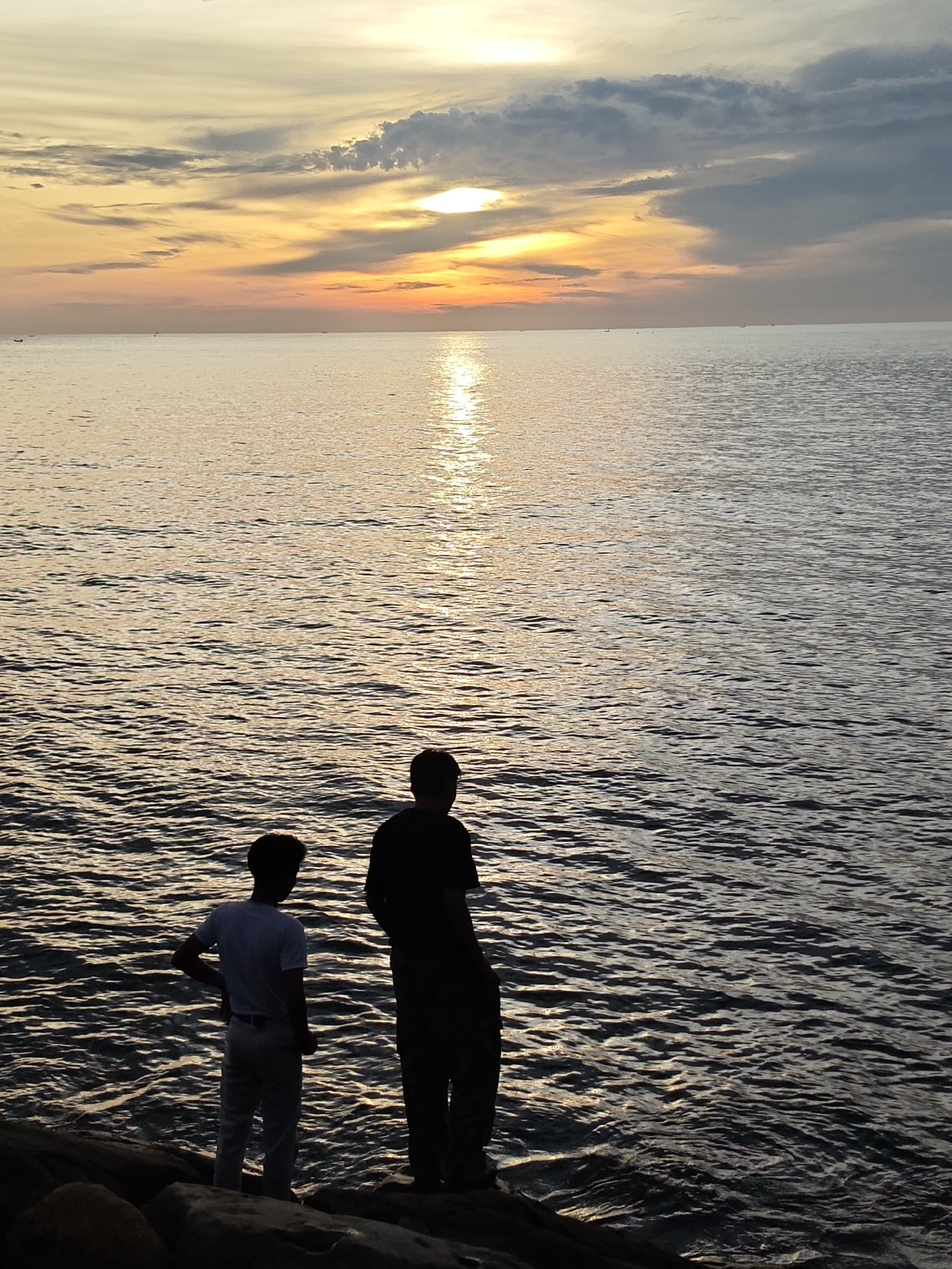 This image displays a coastal scene during a low sun event, characteristic of either sunrise or sunset, in Lhokseumawe, Indonesia. The upper portion of the frame features a sky transitioning from vibrant orange and yellow hues at the horizon to lighter tones and patches of dark grey stratus clouds further overhead, particularly prominent in the upper right and left. The sun, partially concealed by clouds, is visible as an intense bright spot in the upper right quadrant, casting a distinct, shimmering column of light across the ocean surface. The sea, occupying the mid-ground, exhibits a rippled texture reflecting the sky's warm colors, with the sun's reflection creating a golden path extending towards the viewer on the right side of the water. Several small, dark shapes, consistent with distant marine vessels, are faintly visible along the horizon line. In the lower foreground, two male figures are silhouetted against the illuminated water, standing on dark, uneven rocks along the shoreline. The individual on the left is dressed in lighter clothing, with his left hand on his hip, facing towards the sea. The individual on the right, appearing taller and clad in dark attire, is positioned with his back partially to the viewer, also observing the ocean.