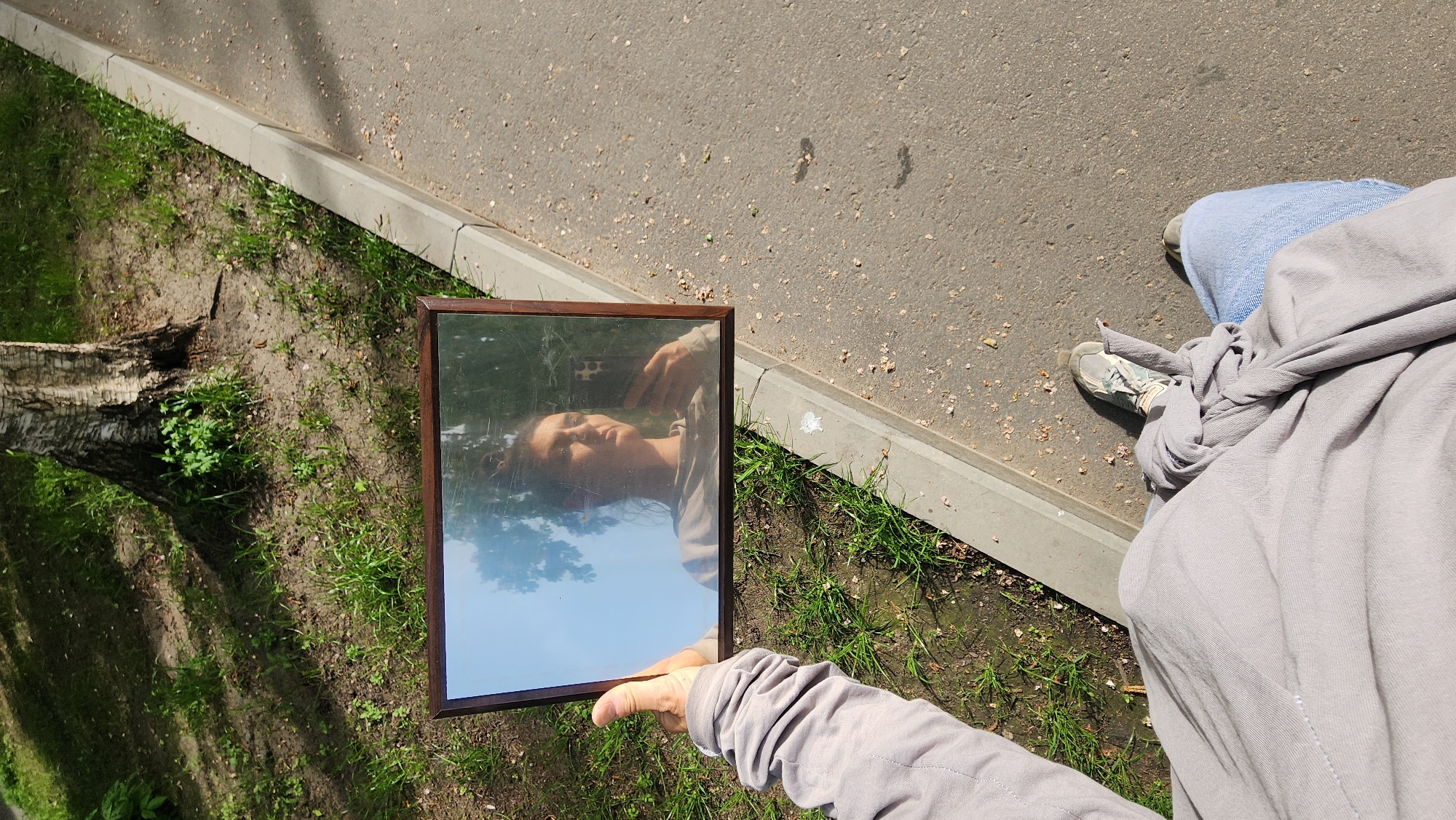 A person holds a square mirror, reflecting themselves and the sky, next to a tree stump and a road in what appears to be Moscow, Russia. The scene is outdoors on a sunny day, with grass growing near the road. The person is wearing light-colored jeans, a gray jacket tied around their waist, and light-colored sneakers. The mirror has a dark brown frame.