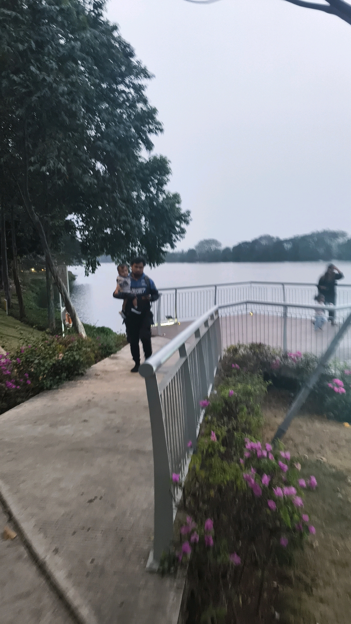 A man is walking on a concrete path with a baby in his arms. He is wearing a black jacket with blue accents and black pants. The man is walking towards the right of the frame. There is a railing on his right, and a body of water on his left. Behind the railing, there is a paved area with a few people standing. In the background, there is a large body of water and trees. The sky is overcast. The scene appears to be a park or recreational area.