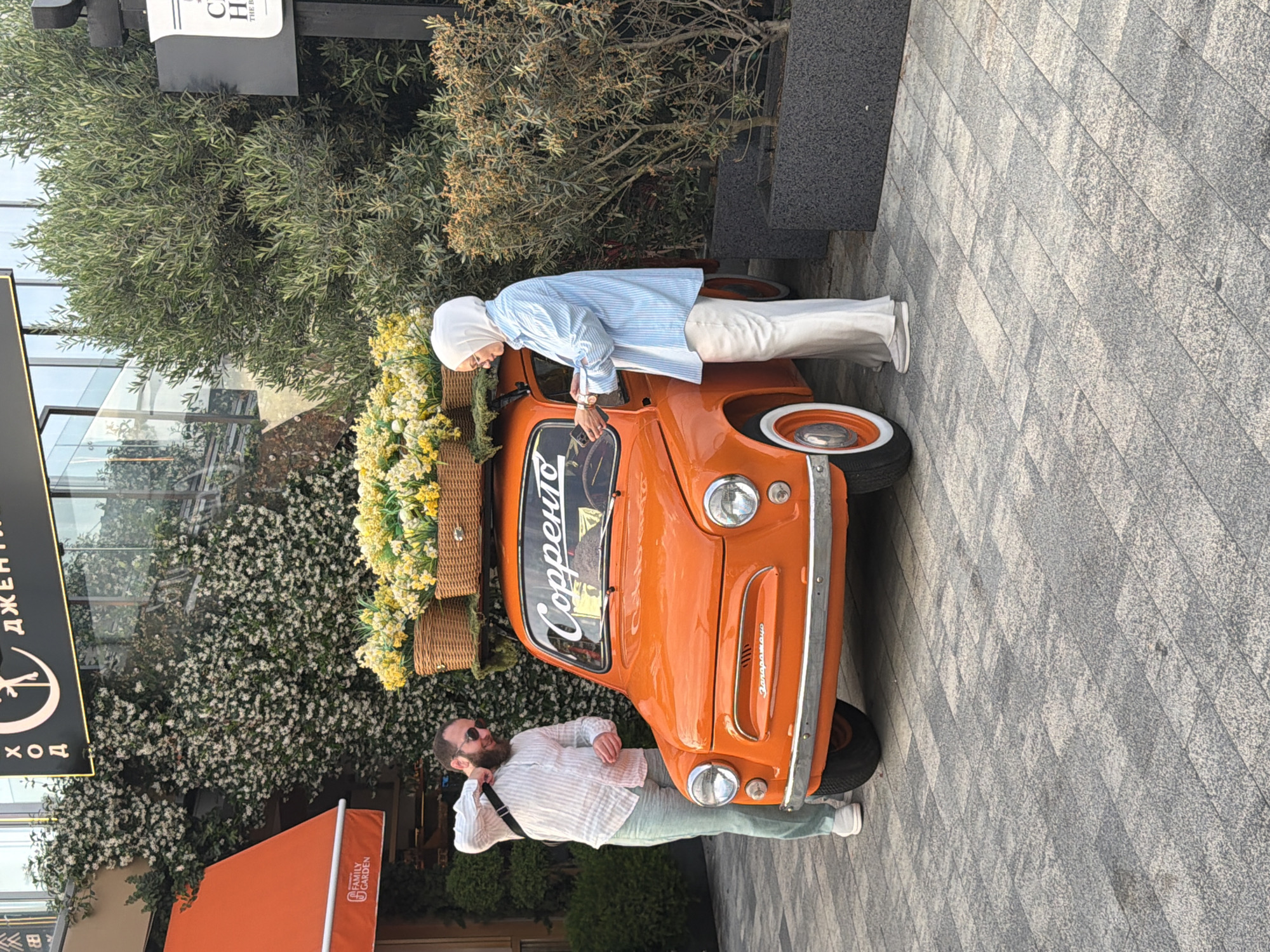 In Sochi, Russia, in front of a building with a facade adorned with white flowering vines, a small orange vintage car is parked on a grey patterned paved surface. The car's roof is decorated with two wicker baskets filled with yellow and white flowers. A man with a beard, wearing a striped white shirt, light green pants, sunglasses, and sneakers, stands to the left of the car, adjusting his bag strap over his shoulder. A woman wearing a white hijab, a blue and white striped long-sleeved shirt, and white wide-leg pants stands to the right of the car, looking at her phone. The background includes a building with an orange awning on the left and large green olive trees in pots on the right.