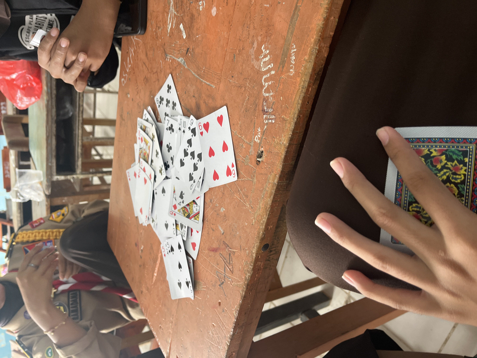 In Pompanua, Indonesia, a group of individuals are gathered around a wooden table, playing cards. The table is cluttered with playing cards, some face up, revealing suits like hearts and spades, and others partially obscured. The individuals are dressed casually, with one person wearing a scout uniform. Their hands are visible, interacting with the cards, suggesting a game is in progress. The environment appears to be an indoor setting, possibly a classroom or community space, with wooden chairs and tables visible in the background. The lighting suggests it is daytime.