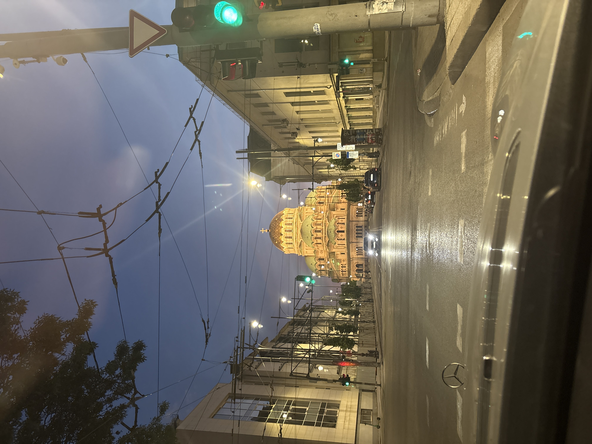 An exterior, eye-level, wide shot captured from a vehicle shows the Alexander Nevsky Cathedral in Sofia, Bulgaria, at dusk. The cathedral, a prominent landmark with a large golden dome and green cupolas, is illuminated and stands at the end of a wide, wet asphalt street. Cars are driving on the street, their headlights reflecting on the wet surface. Traffic lights are visible, with the one on the right showing a green light. Buildings line both sides of the street, and overhead power lines for trams crisscross the sky. Trees and streetlights are also present. The sky is a deep blue, transitioning to darker hues, indicating the end of the day. The foreground shows the hood and windshield of the vehicle from which the photo is taken, with a Mercedes-Benz emblem visible on the hood.