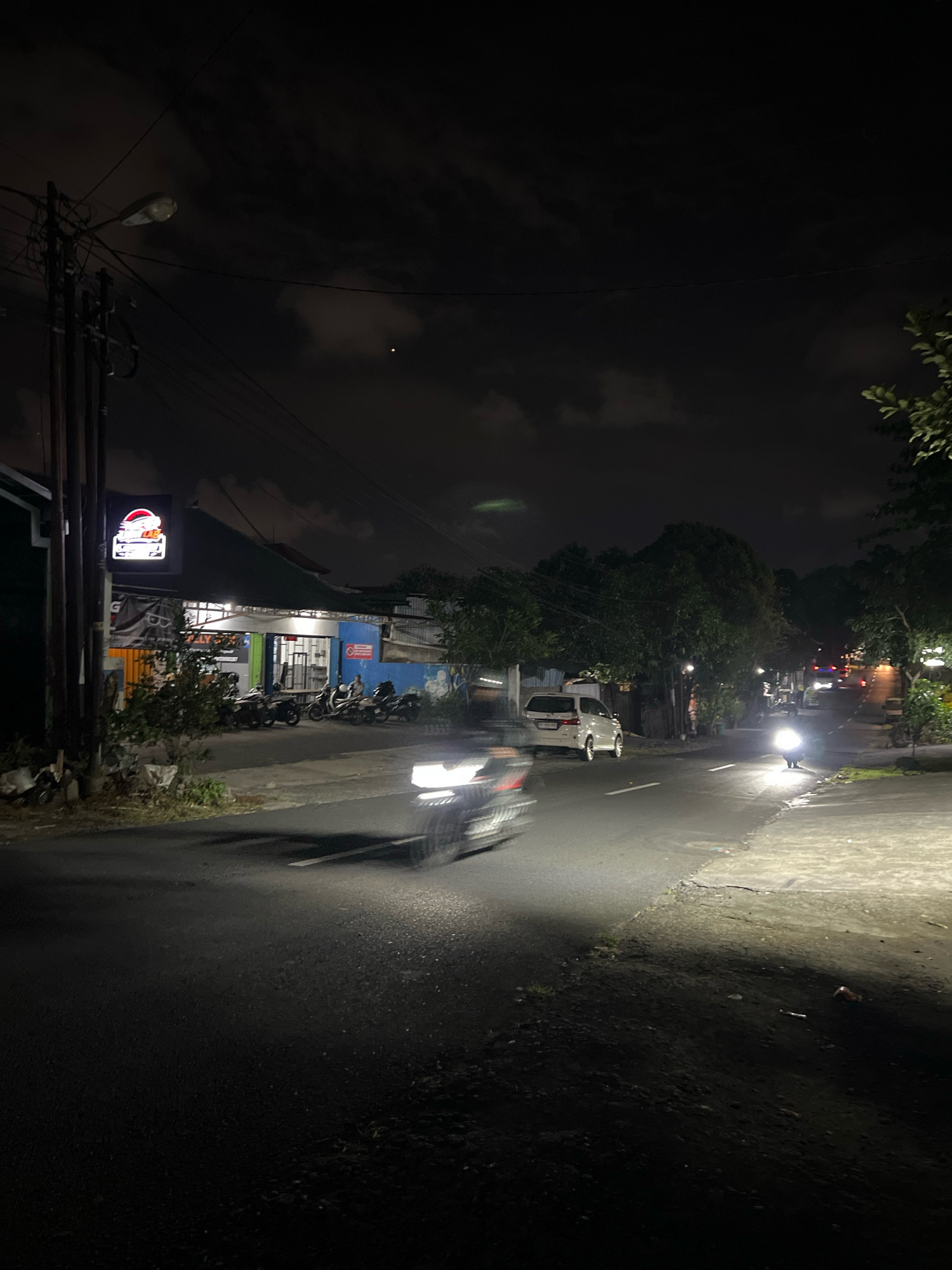 This is a night-time photograph taken on a street in Denpasar, Indonesia. The scene is dimly lit by streetlights and the headlights of passing vehicles. In the foreground, a motorcycle is captured in motion blur, indicating it is moving forward. To the left, a building with a prominent illuminated sign is visible, featuring a red and white graphic of a stylized helmet. Several parked motorcycles are lined up in front of this building. Further down the street, a white van is parked on the right side. The road appears to be asphalt, with a concrete sidewalk on the right. Trees and power lines are also present, contributing to the urban streetscape. The sky is dark with some visible clouds, and a faint light in the sky might be a star or a distant light source. The overall impression is of a quiet, ordinary street scene at night in a Southeast Asian city.