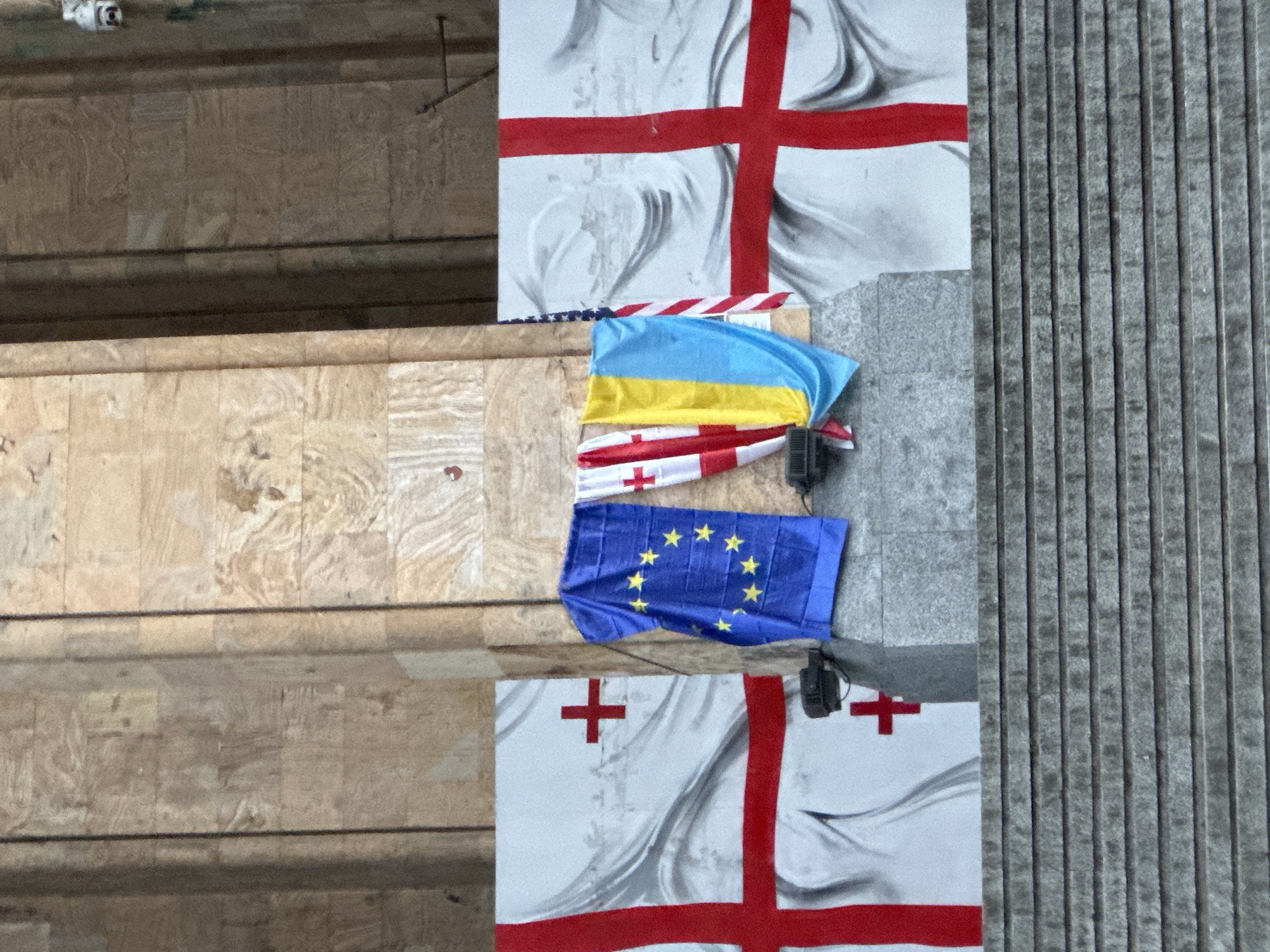 The image shows a stack of flags (Ukraine, Georgia, and the European Union) placed on a ledge or platform in Tbilisi, Georgia. Georgian flag imagery is also visible above and below the other three. A security camera is also present above the scene. The background is composed of wall surfaces, including a corrugated grey wall, and the wooden ledge where the flags rest.