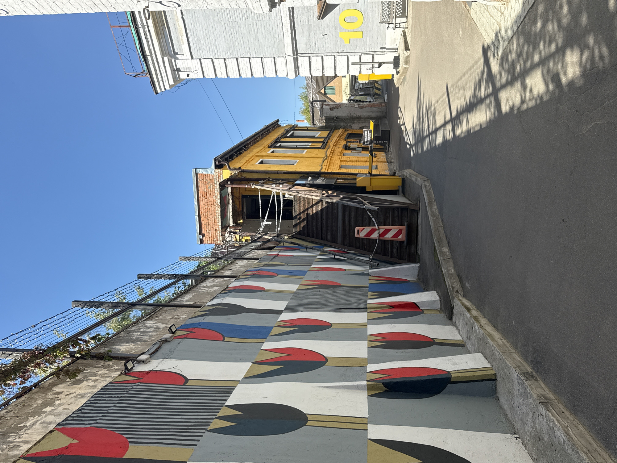 The image shows a narrow street in Moscow, Russia, with a large mural on the left wall. The mural depicts stylized tulips in red, black, and blue against a grey and white background, arranged in a repeating pattern. The wall is topped with a fence with barbed wire. To the right of the mural, there is a steep set of outdoor wooden stairs leading up to a yellow building with a brick facade. The yellow building has several windows and a balcony. Further down the street, there are more buildings, including a white brick building with the number "10" painted in yellow. Shadows of tree branches are cast across the asphalt road. The sky is clear and blue. The scene appears to be an alleyway or service entrance in an urban area.