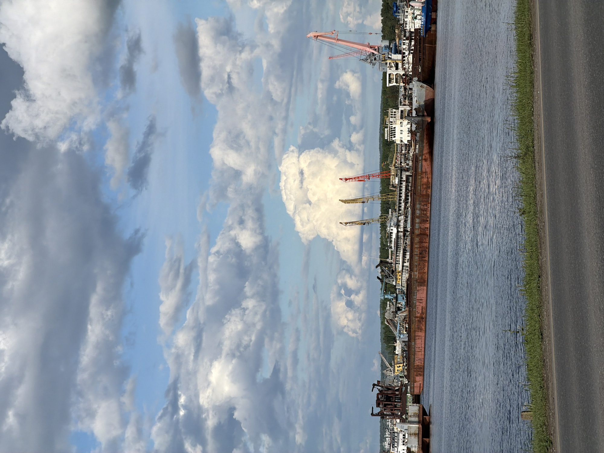 The image depicts a river scene alongside a road, under a sky filled with clouds. A row of docked barges and cranes lines the riverbank. The location is Chaikovsky, Russia, based on the type of equipment. The perspective is vertical with the sky filling the majority of the frame. A road borders the other side of the river bank.