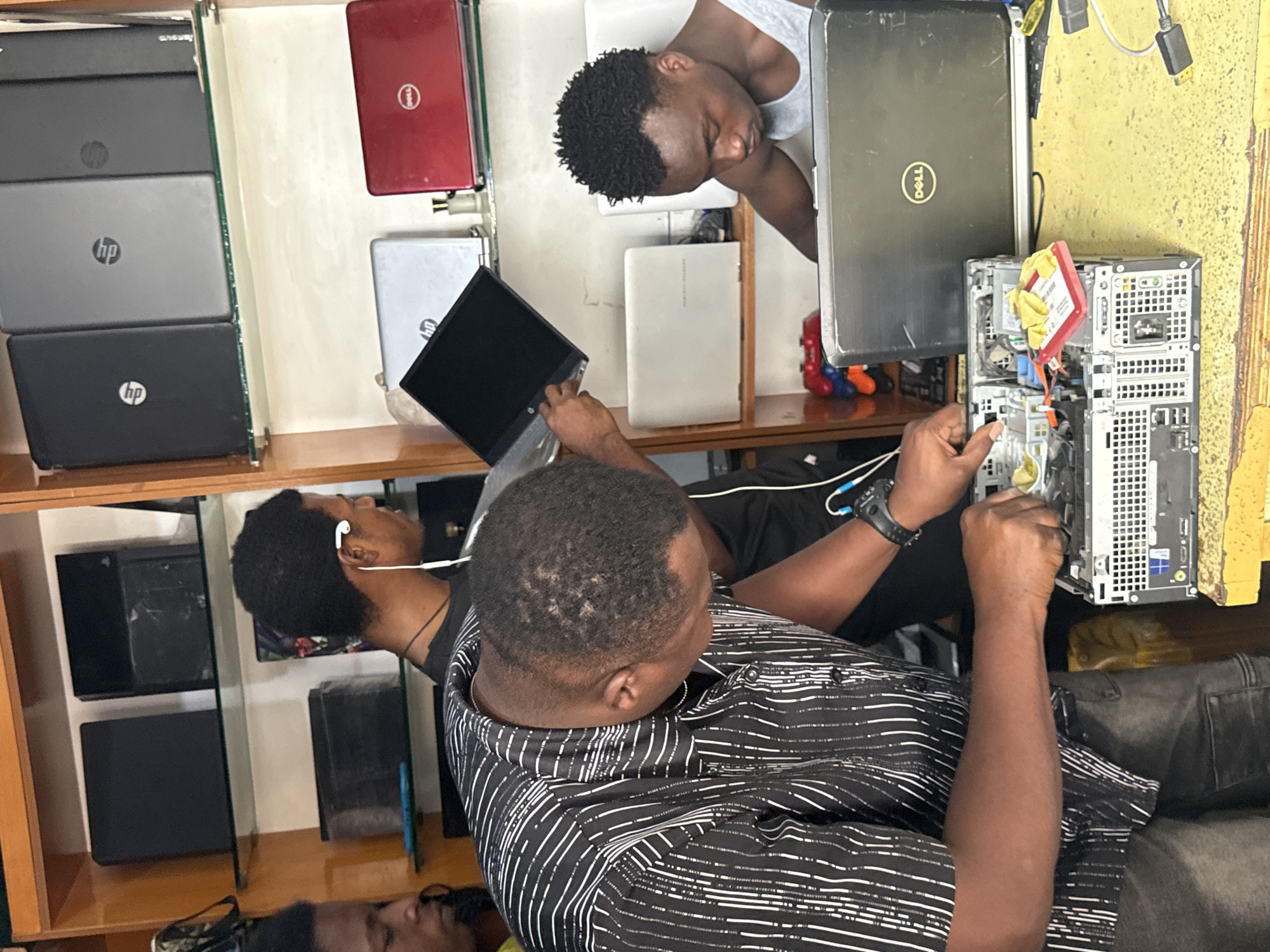 The image depicts a computer repair shop located in Lekki, Nigeria. In the foreground, a man wearing a black and white striped shirt is focused on repairing a computer tower, with his hands engaged in the internal components. Next to him, another man wearing a grey tank top is also working on computer equipment. The background shows shelves filled with various electronic devices, including laptops, computer towers, and what appear to be gaming consoles. The environment is typical of a busy retail or service establishment in the area, with equipment and supplies visible. The lighting suggests an indoor setting, likely during daytime.