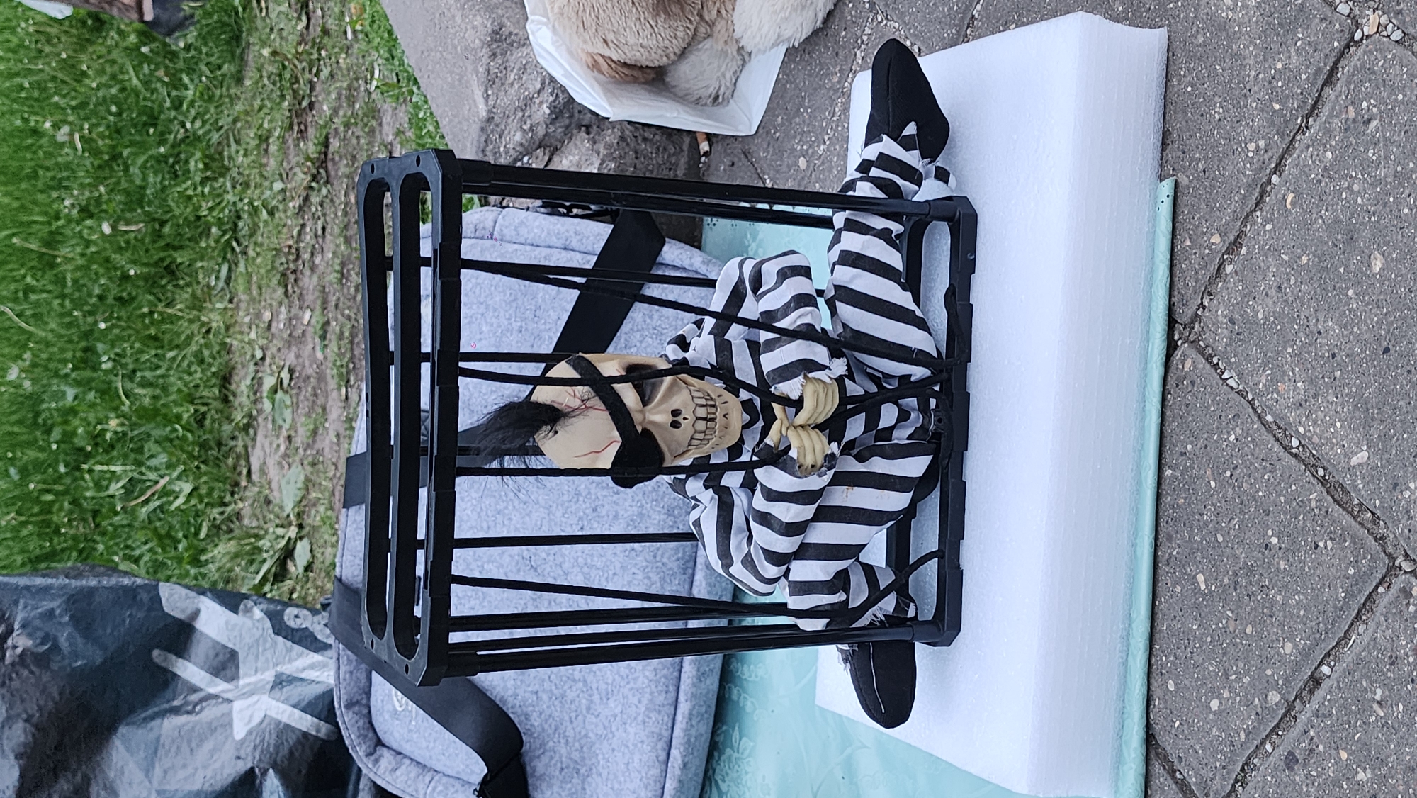 The image depicts a Halloween decoration consisting of a skeletal figure dressed in a black and white striped prisoner uniform, contained within a black metal cage. The skeleton has a black eye patch and is bound with black rope. The cage sits on top of a white rectangular block, possibly styrofoam. The background includes a patch of green grass, a grey tarp-like material, and a paved area with grey bricks. There is also a pet bed with fur inside. There is no information in the image to suggest it is in Moscow, Russia.