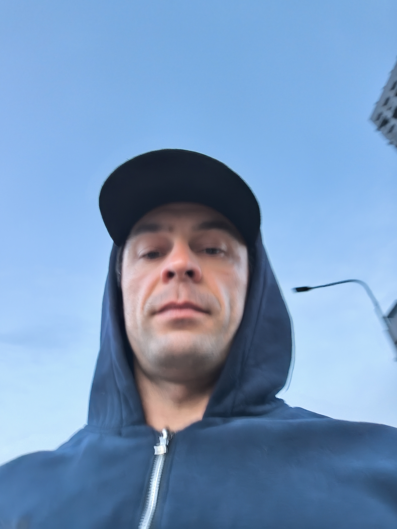A selfie photograph features a man wearing a dark blue hooded sweatshirt and a black baseball cap, positioned outdoors against a bright blue sky. He appears to be standing at a lower angle, capturing the image upwards towards his face. In the background, a street lamp and a corner of a tall building are visible, suggesting an urban location. Considering the style and subject, the location cannot be verified as Moscow, Russia without further evidence.