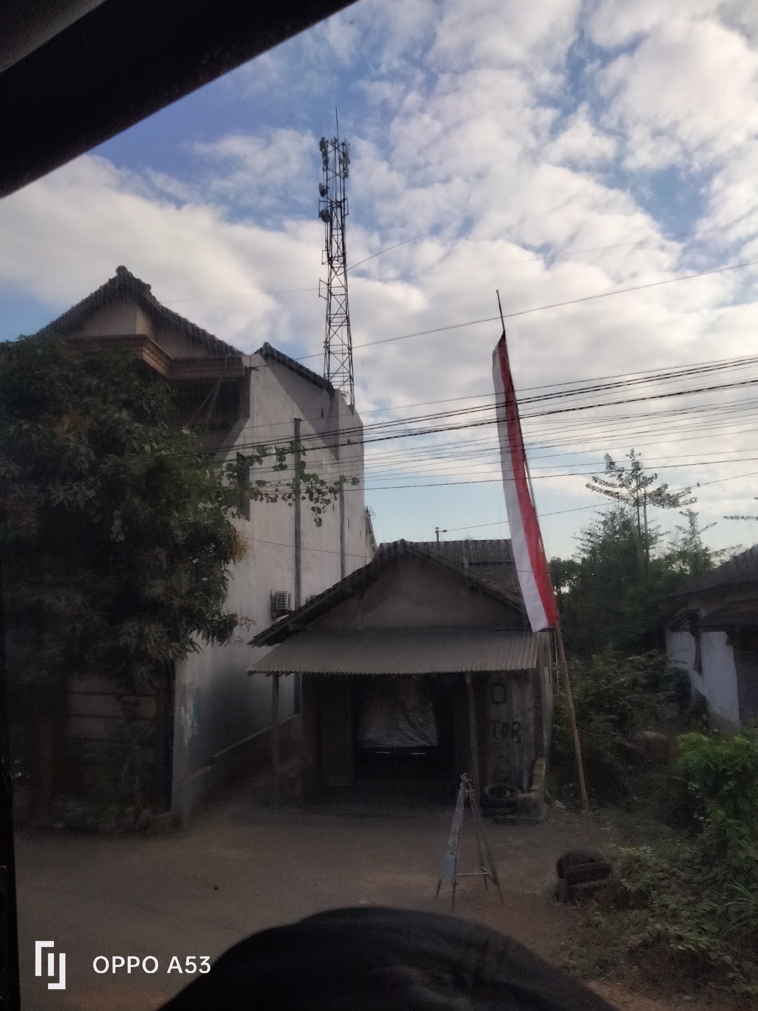 The image captures a street scene in Buntaran, Indonesia, under a cloudy sky. In the foreground, a dark, out-of-focus object, likely part of a vehicle or obstruction, occupies the bottom portion. A logo indicating "OPPO A53" is visible in the lower left corner. The environment includes a dirt road, a white building with a dark roof and a large tree to its left, and a smaller, weathered structure with a corrugated metal roof in front of it. A tall cell tower stands prominently in the background. An Indonesian flag, red and white, is planted near the smaller structure, and electrical wires crisscross the sky. Dense green foliage is present on the right side of the image, with more buildings visible in the distance. The overall impression is of a typical residential or mixed-use area in a suburban or rural setting.