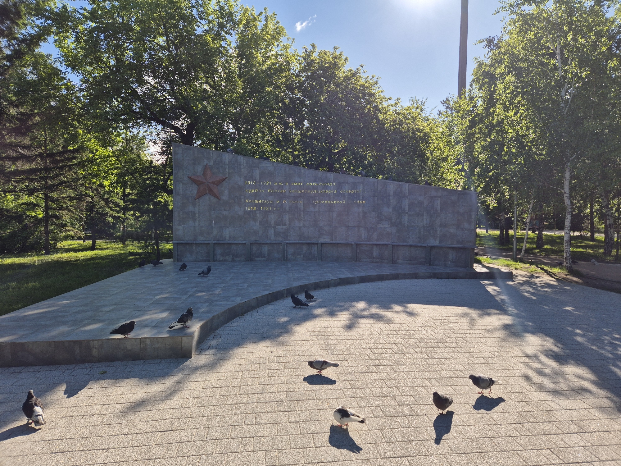 The image depicts a memorial in a park. The memorial is a large, gray stone slab with a red star on the upper left. The slab is inscribed with text in two languages. In front of the slab is a paved plaza where several pigeons are scattered. The plaza is made of gray paving stones, and the light casts shadows from the surrounding trees onto the stones. The background features lush green trees and grass, suggesting a well-maintained park setting. The overall atmosphere is tranquil. The location is in the city of Unknown, Unknown.