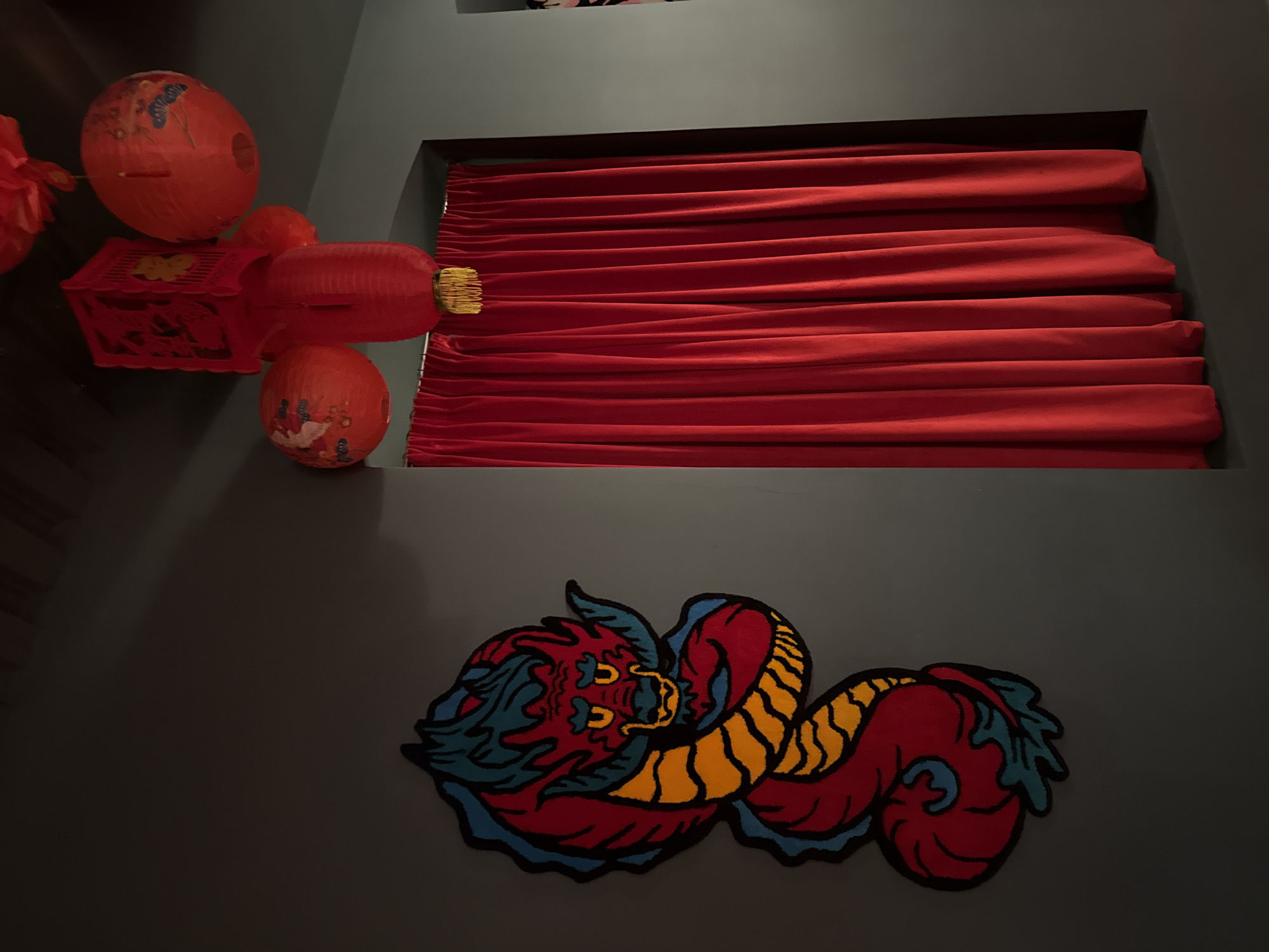 The image displays an indoor space in Moscow, characterized by dim lighting and an East Asian-inspired decor theme. The walls are a muted dark grey or teal, setting a somber backdrop for the vibrant red decorations.
On the left side of the frame, several red Chinese-style lanterns are arranged. These include a large, spherical lantern adorned with painted blue butterfly-like motifs, and a rectangular, intricately carved lantern. Adjacent is a cylindrical, accordion-pleated lantern featuring a distinctive golden tassel. Below these, a smaller spherical lantern is visible, with a painted design that appears to be a bird or floral pattern.
In the upper-right, a recessed opening, possibly a window, is filled with rich, red pleated curtains or blinds. These are drawn horizontally, creating distinct folds and adding depth to the red color scheme.
Dominating the lower wall is a large, colorful dragon motif, which appears to be an embroidered or appliquéd piece. The dragon is depicted with a fierce expression, primarily in red with bold yellow stripes on its body, blue accents for its mane and frills, and strong black outlines.
The overall scene conveys a festive or culturally themed atmosphere, likely in a restaurant or specialized venue within Moscow. The artificial lighting creates a warm and inviting ambiance. No people are visible, and there is no discernible text in the image.