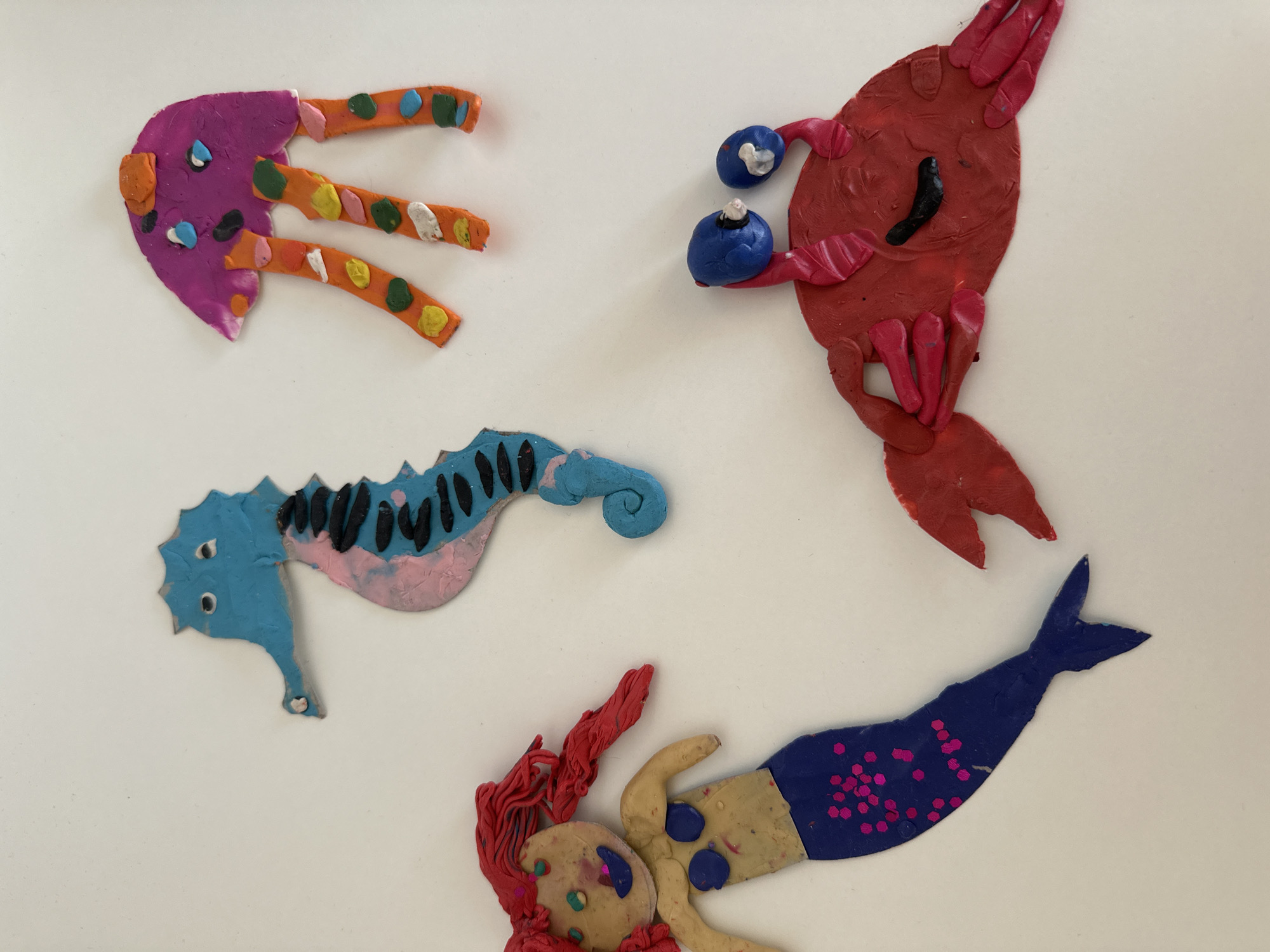 The image shows four figures made of modeling clay against a white background.
The figures appear to represent marine life: a jellyfish, a lobster or crab, a seahorse, and a mermaid.
The jellyfish is purple with orange tentacles decorated with colorful dots.
The lobster is red with large blue eyes.
The seahorse is blue and pink with black markings on its back.
The mermaid has red hair, a tan body, and a dark blue tail with pink dots.
The specific location of Chaikovsky, Russia, cannot be inferred from the image.