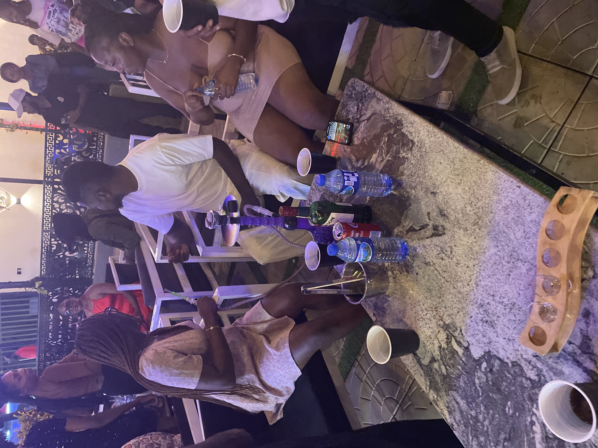 This media file captures an evening social gathering at a lounge or outdoor bar setting in Jos, Nigeria.
**Subjects:** The image features several individuals, both men and women, socializing. In the foreground, a woman with braided hair, wearing a light-colored dress, is seated next to a table. Another woman in a similar dress is seated opposite her, holding a plastic water bottle. A man in a white t-shirt is also seated nearby. More people are visible in the background, standing and seated on tiered structures, dressed in casual evening attire.
**Objects:** A central marble-patterned table holds an assortment of items: multiple plastic water bottles, some bearing the brand "SWARO"; an empty red can with "SON" visible (possibly a soft drink or energy drink); a green bottle partially revealing "JAMESON" (likely whiskey); and several black and white plastic cups. A smartphone displaying a screen is also on the table. An ice bucket with tongs rests beside one of the seated women, and a shisha pipe with multiple hoses is visible near the table. A wooden caddy designed to hold small glasses, with some glasses in place, is also on the table.
**Scene/Setting:** The event takes place in an outdoor or semi-outdoor area, characterized by decorative tiled flooring and artificial grass sections. Metal railings and contemporary white-framed seating structures are present. The artificial lighting, including subtle purple hues, indicates it is nighttime or late evening.
**Activity or Event Taking Place:** The people are engaged in a social gathering, likely a party or relaxed get-together. They are seen relaxing, conversing, and consuming drinks. The presence of alcohol, water, and shisha suggests a leisure activity.
**Notable Details:** The ambient lighting creates a relaxed and social atmosphere. Attire is casual, suggesting comfort for an evening out. The overall setup points to a modern entertainment venue. The individuals appear to be enjoying the event.
**Visible Text:** "SWARO" is clearly visible on the labels of the plastic water bottles. "JAMESON" is partially visible on the green bottle, and "SON" is seen on the red can.