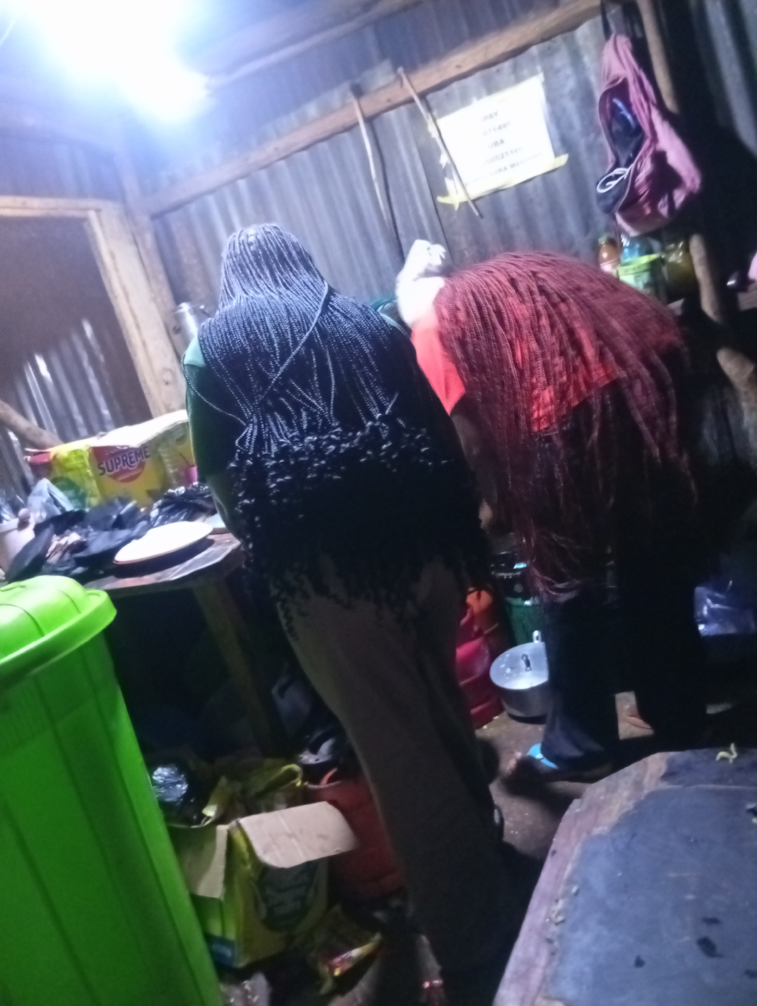 The image depicts an indoor scene, possibly a market stall or a workshop, in an urban environment. Two individuals are the primary subjects. One person, facing away from the camera, has long black braided hair that falls down their back. They are wearing a green garment. Another person, partially obscured, has long, voluminous red hair and is seen bending over.
The background is rustic, with corrugated metal walls. Several objects are visible on a wooden surface, including a white plate, a yellow package labeled "SUPREME," and other unidentifiable items. A bright overhead light illuminates the scene from the upper left. In the foreground, a large green plastic bin is prominent on the left side of the image. A pink bag hangs on the wall in the background, along with a sign that appears to display payment information. The floor is dirt or concrete. The overall impression is one of a busy, functional space, likely associated with commerce or a craft.