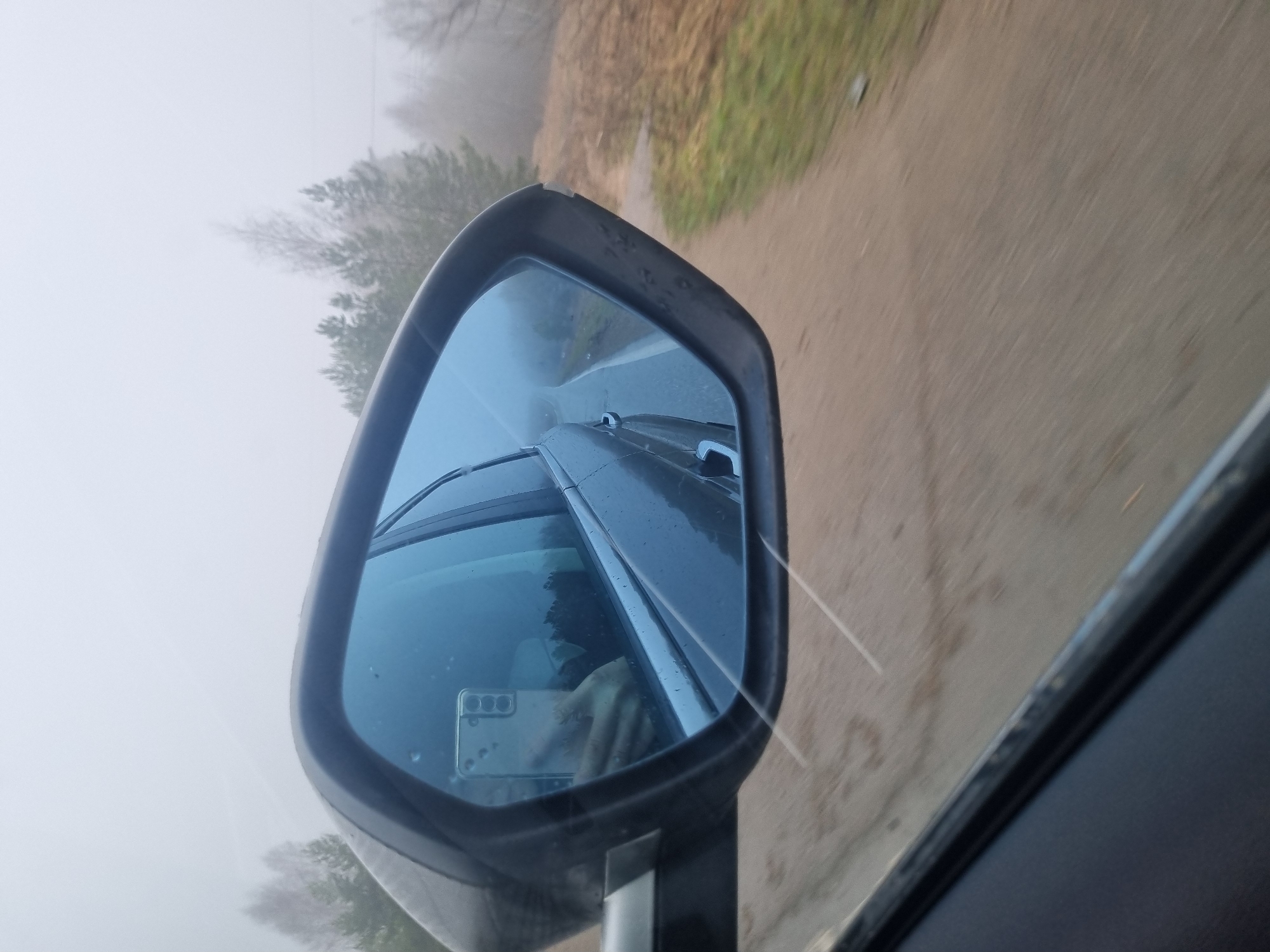 The image shows the side mirror of a car, reflecting the car's body, windshield wipers, and the driver's hand holding a smartphone. The background visible in the mirror is foggy. The road outside the car is blurred, indicating motion. Trees are visible along the road, obscured by fog. It is not possible to determine the location in the city of Chaikovsky, Russia from this image.