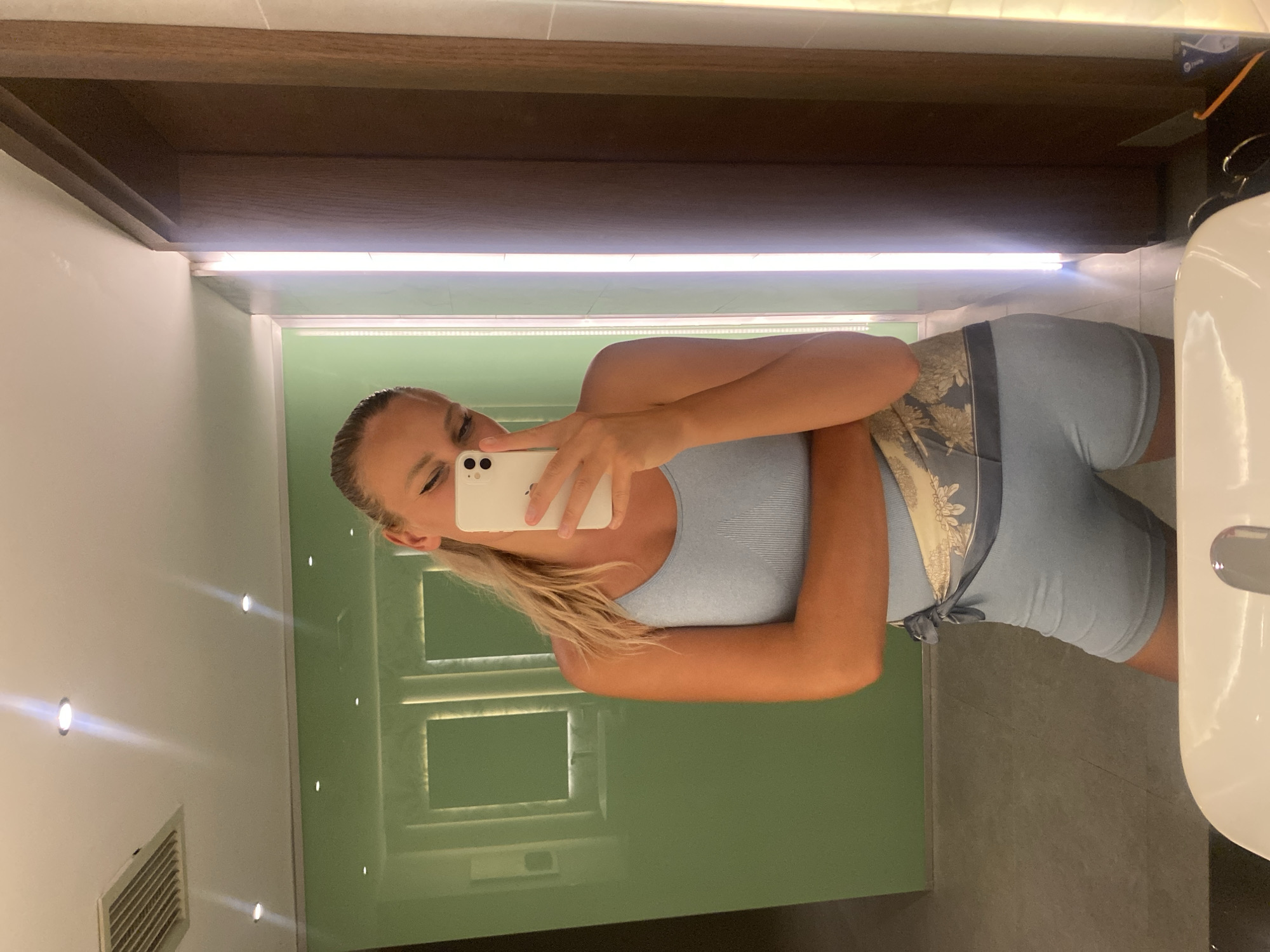 A young woman with blonde hair pulled back into a ponytail takes a selfie in a bathroom mirror. She is wearing a light blue tank top and matching light blue shorts, with a patterned sash tied around her waist. Her left arm is bent and her hand is resting on her hip, while her right hand holds the phone to take the picture. The bathroom features a light green wall behind her, a white sink in the foreground, and a dark wood frame around the mirror. The lighting is bright, with spotlights visible on the ceiling and a vertical light strip on the right side of the mirror. The location is not specified in the image, and therefore cannot be identified as Aksu, Turkey.