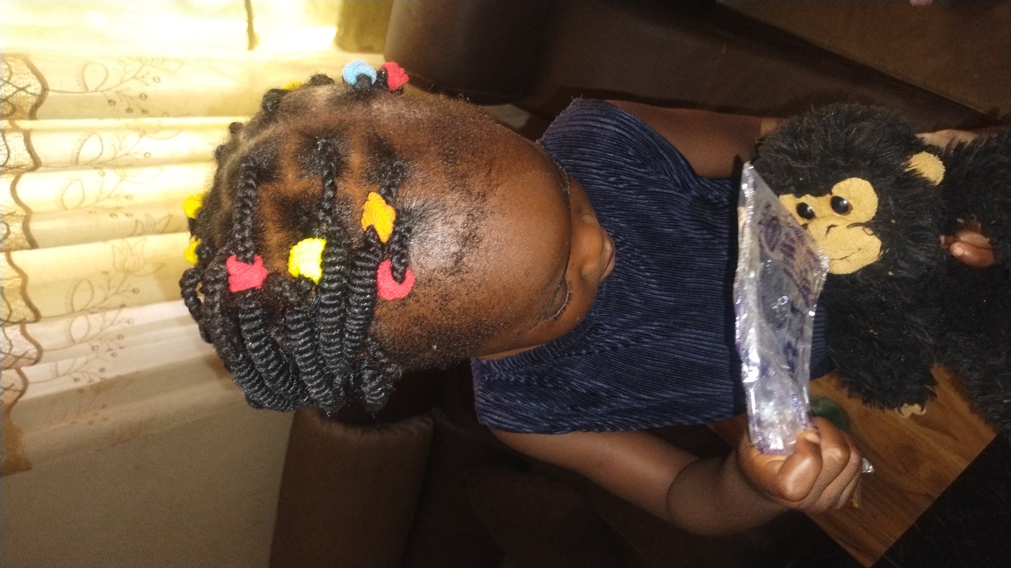 A young Black girl with braided hair adorned with colorful beads sits on a sofa, holding a plastic-wrapped item and a stuffed monkey. The girl is wearing a navy blue pleated sleeveless top. The background shows curtains and a sofa. The scene appears to be indoors, likely in a residential setting in Jos, Nigeria.