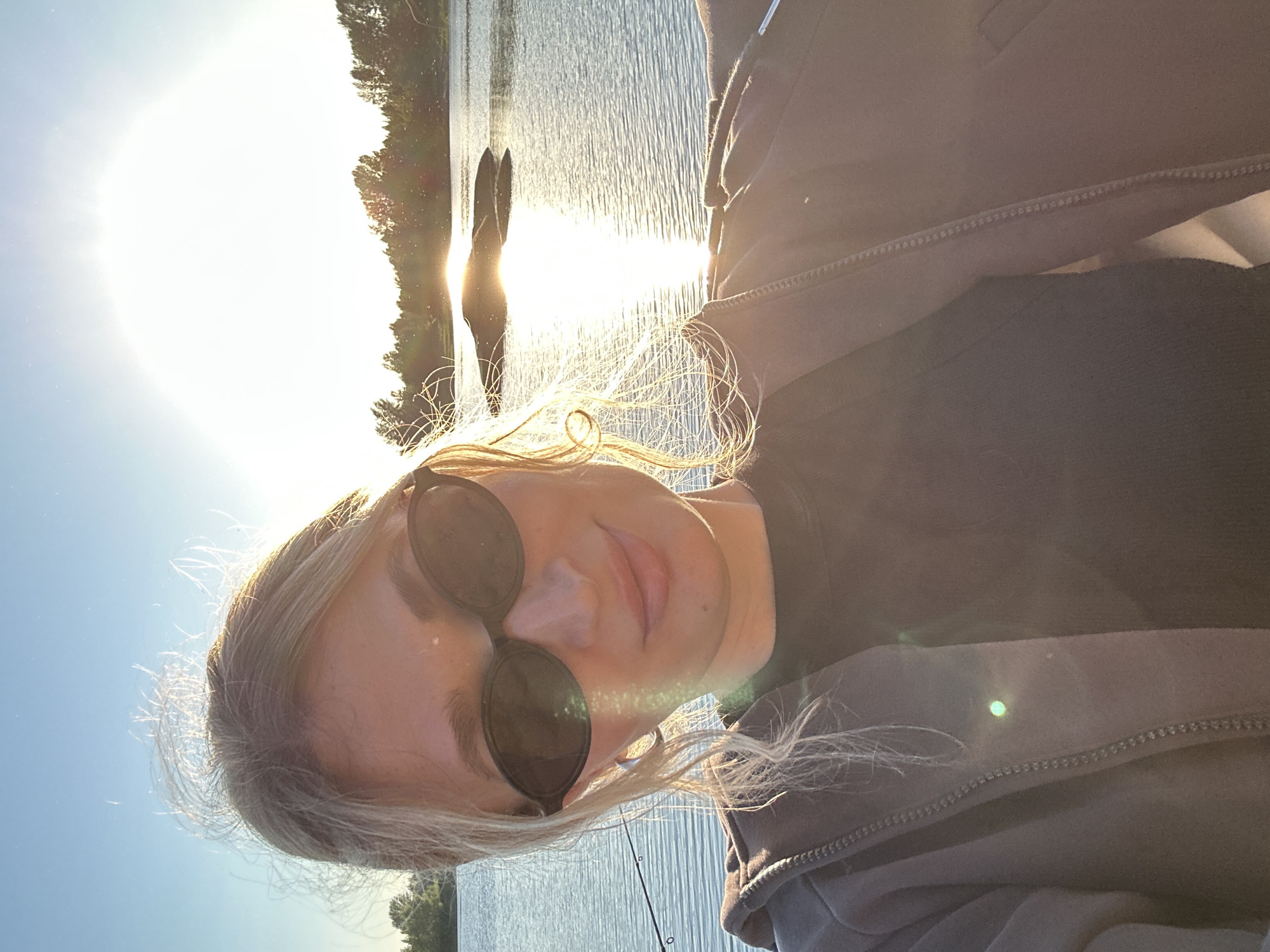 A medium shot shows a young woman with blonde hair and sunglasses taking a selfie outdoors by a body of water. She is wearing a dark t-shirt and a brown zip-up hoodie. The sun is setting, casting a warm glow on the scene and creating a lens flare in the upper part of the image. The water is calm, with a few ripples and reflections of the sky. In the background, a line of trees is visible on the horizon. The setting appears to be a natural landscape, possibly a park or shoreline, with a fishing rod visible to the left of the frame. The overall mood is relaxed and pleasant, with the golden hour light contributing to a serene atmosphere.
