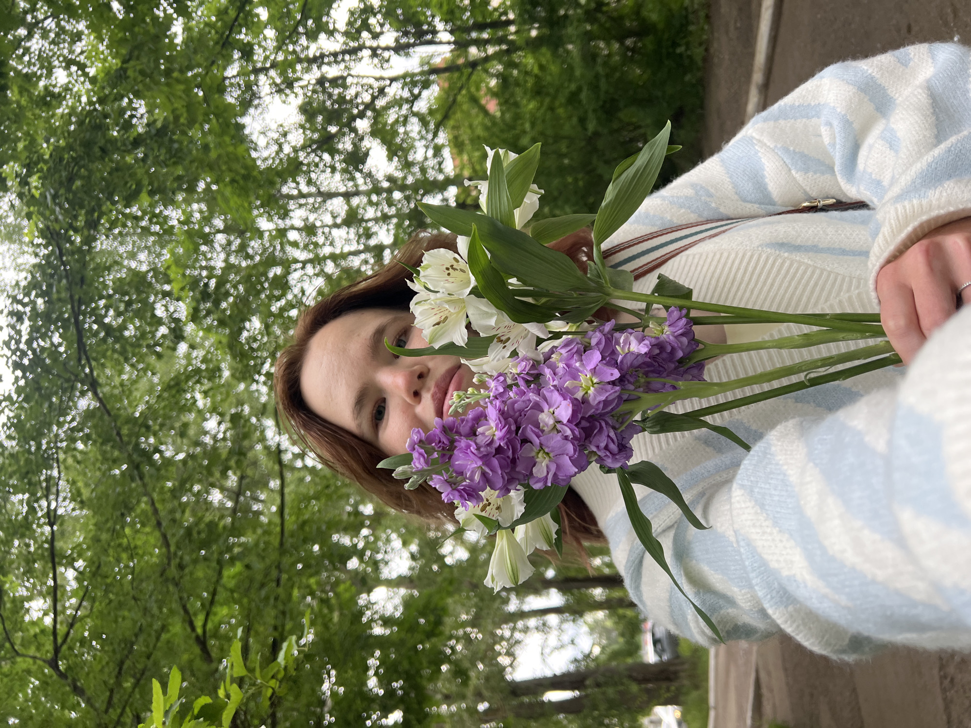 The image is a close-up, sideways-oriented portrait of a young person, likely female, captured from a low angle. She has light brown hair and a serene, neutral expression, with her gaze directed towards the viewer. She is holding a vibrant bouquet of flowers close to her face, consisting of white alstroemeria with speckled green centers and stalks of fragrant purple stock (Matthiola incana).
The subject is wearing a cozy, knitted sweater with a cream or off-white base, adorned with light blue wavy or patterned stripes. A dark, possibly leather, strap from a bag or accessory is visible over her left shoulder. A delicate ring is worn on the ring finger of her left hand, which is gently cradling the flowers.
The background is dominated by a dense canopy of lush green trees, suggesting an outdoor setting such as a park or a tree-lined street. The bright, soft light filtering through the leaves indicates it is daytime, possibly overcast or late afternoon, as there are no harsh shadows. A hint of a paved surface or pathway is visible in the upper right corner of the image (relative to its original unrotated orientation). The overall mood is calm and natural. No visible text is present in the image. The location context for this scene is Chaikovsky, Russia.