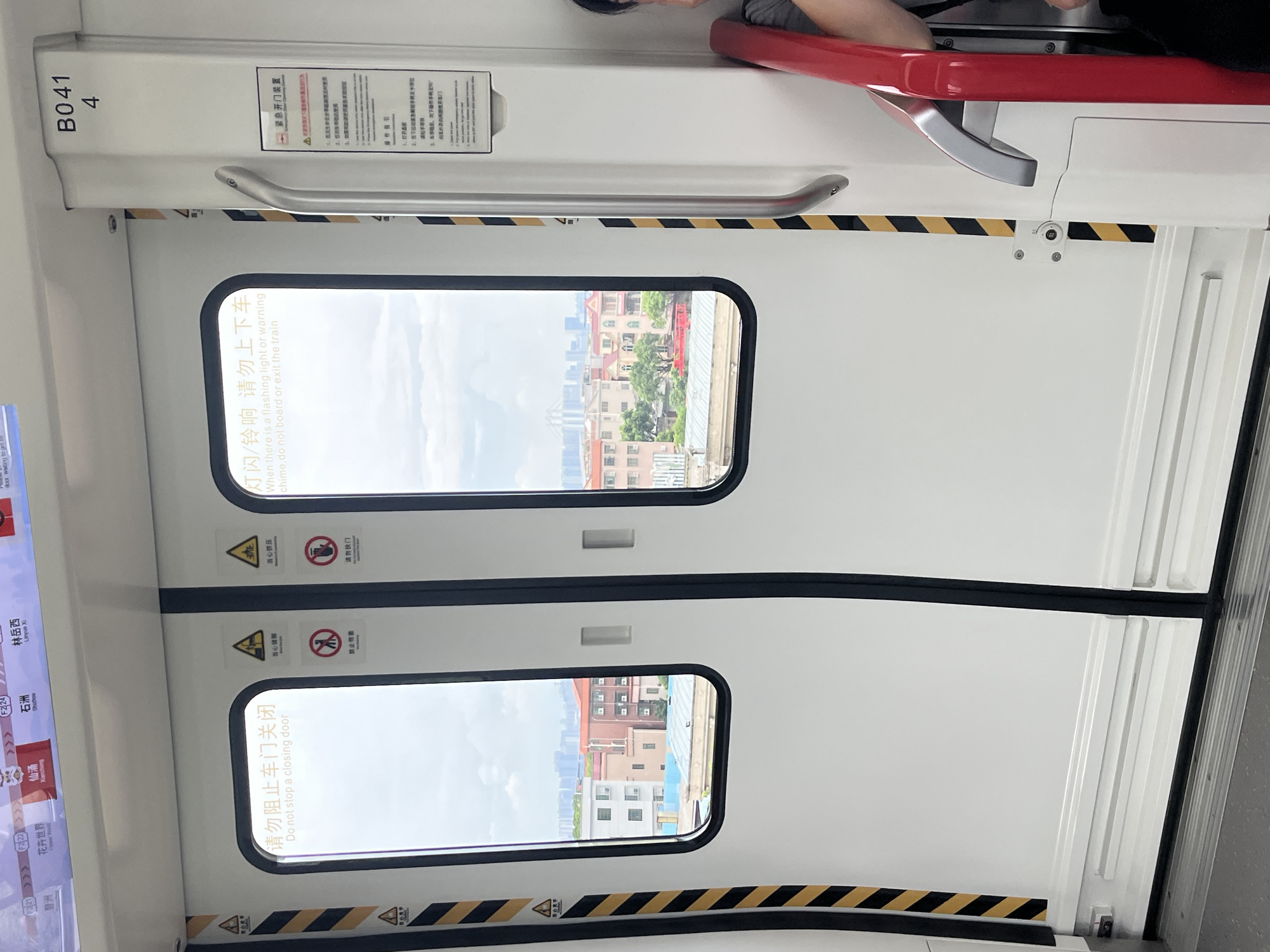The image shows the interior of a subway train carriage, likely in Shunde District, China, based on the visible station names on the overhead display (e.g., 杏坛 Shunde, 仙涌 Xianchong, 石洲 Shizhou, 林岳西 Linyuexi). Two sets of white subway doors are visible, with one slightly ajar. Yellow and black hazard stripes run vertically along the door frame. Various warning signs are affixed to the doors, including a sign in Chinese and English instructing passengers not to block closing doors and symbols indicating no smoking and no touching. The right door also has an emergency door opening device notice. Through the windows of the doors, a city landscape with buildings and some trees can be seen under a cloudy sky. A metal handrail is attached to the side of the carriage, and a red pole is present in the foreground. The number "B041 4" is printed on the frame of the right door. The floor of the train is grey and ribbed. A portion of a person's arm and shoulder, wearing a dark top, is visible on the right side of the image, holding onto the red pole.
