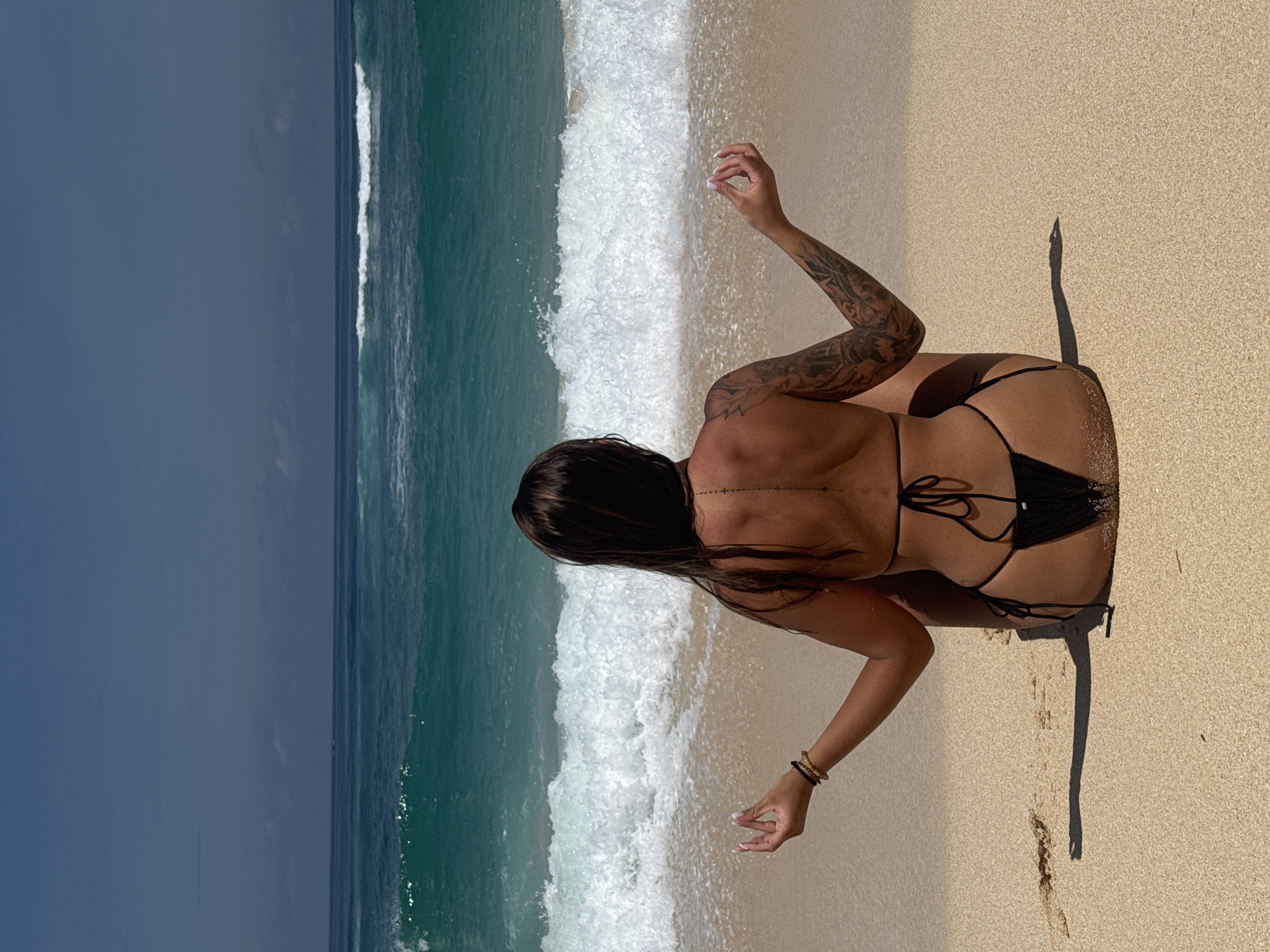A woman is on a sandy beach in Kuta Selatan, Indonesia. The woman, who has dark hair, dark skin, and tattoos on her upper arms, is wearing a black bikini. She is crouched with her back to the camera and her arms extended. The beach is bordered by the ocean, which has turquoise water and white waves. The sky is dark.