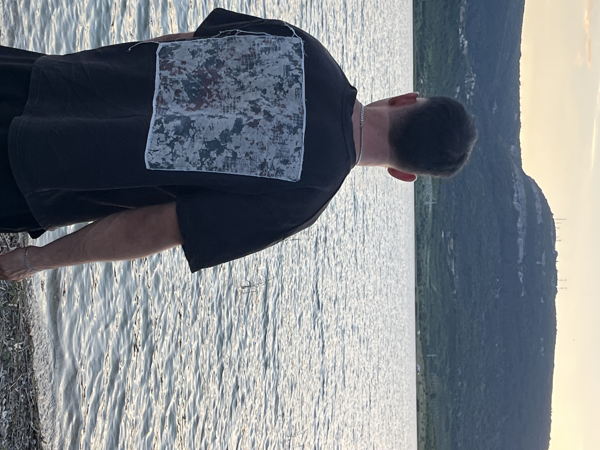 A person stands on a pebble-strewn shore, facing away from the camera and looking out over a body of water towards a large, tree-covered hill under a pale sky. The person is wearing a dark t-shirt with a rectangular graphic on the back, featuring a distressed, textured design with shades of gray, white, and some dark red or brown. They are also wearing dark shorts and a silver chain necklace. The water is rippling, and sparse vegetation grows along the shoreline. The hill in the background has buildings and communication towers visible on its crest, and a small village is nestled on its lower slopes. The overall scene suggests a natural landscape near a settlement, possibly on the outskirts of Shumen, Bulgaria, given the prominent geographical features.