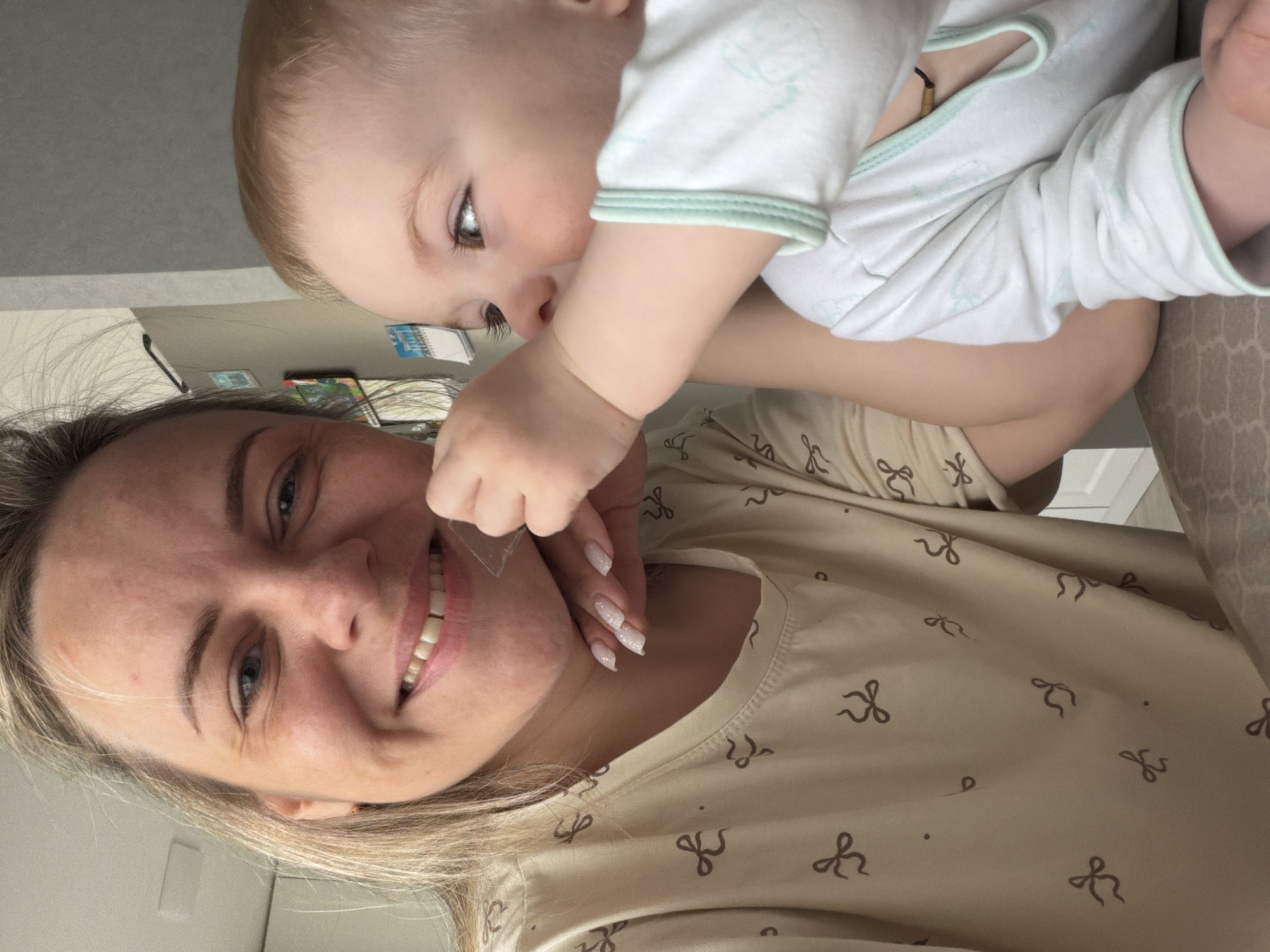 This vertically oriented, close-up photograph captures a tender and joyful interaction between a woman and a baby in an indoor setting. The image is composed from a low angle, possibly a selfie, looking up towards the baby.
The woman, likely the mother, occupies the lower portion of the frame. She has light blonde hair, fair skin, and blue eyes, and is seen smiling broadly, revealing her teeth. Her long fingernails are manicured with light, possibly glittery, polish. She wears a light-colored top patterned with small, dark brown bow ties.
Above her, a baby with light blonde hair and fair skin gazes downwards, reaching out with one hand towards the woman's face or chin. The baby is dressed in a white long-sleeved onesie with light green trim around the neckline and cuffs. A dark string, possibly a necklace, is visible around the baby's neck. Both subjects appear happy and engaged, conveying a warm and affectionate atmosphere.
The background is softly blurred but reveals hints of an indoor environment, including a light-colored wall or furniture and a partial view of colorful objects, possibly books or magazines, further back. The lighting is soft and even, suggesting daytime or a well-lit interior. There is no legible text visible in the image. While the location context provided is Votkinsk, Russia, the visual content does not offer specific geographical cues beyond a typical home interior.