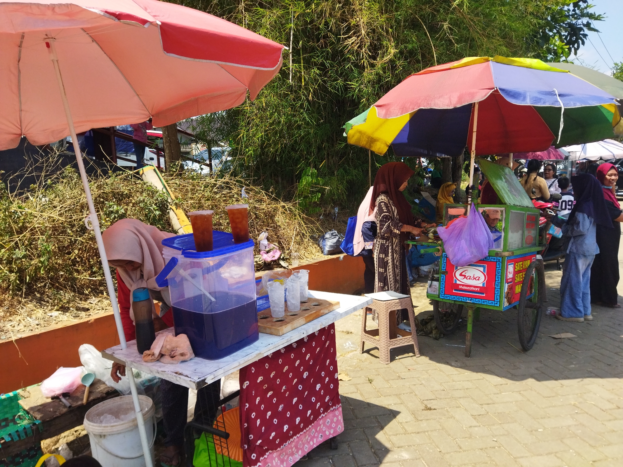 This is a street scene in Semarang, Indonesia, on a sunny day. Two stalls are set up on a paved walkway, shaded by large, colorful umbrellas. The stall on the left features a table covered with a red and white polka-dot cloth. On the table are several cups of iced tea, a large blue plastic container filled with dark purple liquid, and a bottle of dark liquid. A person wearing a light-colored hijab and red shirt is standing behind the table, attending to the drinks. In front of the table is a white bucket filled with ice.
To the right, a food cart is parked, adorned with a colorful umbrella and various labels. People are gathered around this cart. One woman, wearing a dark hijab and a patterned dress, is serving food from the cart. Another woman, in a blue striped shirt and blue pants, is standing nearby. A younger person in a dark shirt with the number "09" is also visible. The background is filled with lush green foliage, likely bamboo, and other vegetation, with a glimpse of parked vehicles suggesting a roadside location. The overall impression is of a vibrant street-side food and beverage vending area.