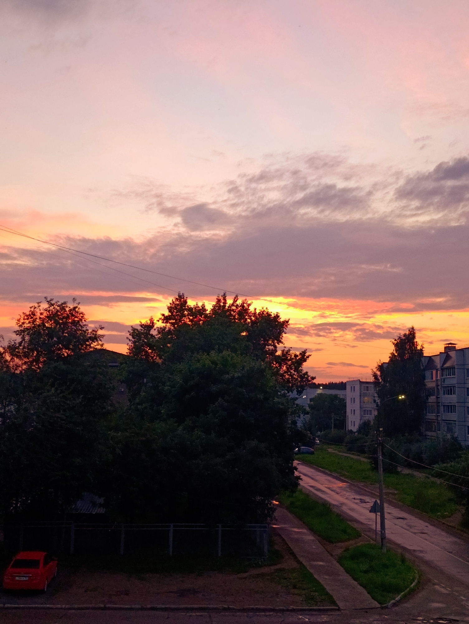 The image captures a sky ablaze with the colors of sunset, transitioning from soft pinks and purples at the top to vibrant oranges and yellows near the horizon. Silhouetted against this dramatic sky are the dark masses of trees, with the fiery sunset peeking through their foliage, creating a striking contrast.
In the foreground, a small red car is parked on a paved surface. A dark metal fence runs along the edge of the property. Behind the trees, the landscape transitions to urban elements. A road, wet and reflecting the twilight hues, curves through the scene, bordered by grassy areas and sidewalks. A utilitarian street lamp stands near the road. Further in the distance, apartment buildings, characteristic of Russian urban architecture, rise against the fading light. The overall impression is that of a residential area in the city of Chaikovsky, Russia, during the magical hour of sunset. There are no people visible in the image.