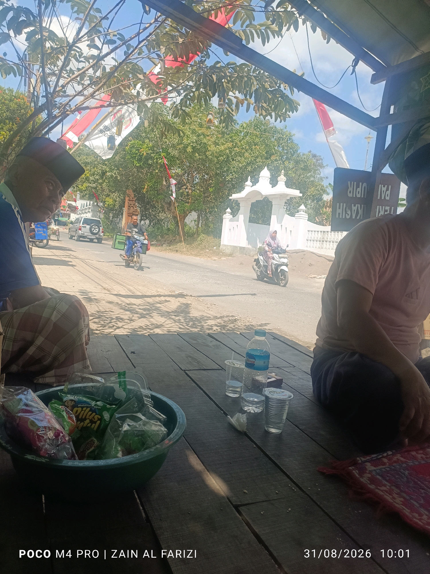 The image captures an outdoor scene in Cendagah, Indonesia, likely a roadside stop on a sunny day. In the foreground, two individuals are seated on a wooden platform, partially shaded by an overhead structure. To the left, an elderly man wearing a dark peci and a blue shirt with a checkered sarong is seated. To the right, another person in a light pink t-shirt is visible from the side. On the wooden surface in front of them, a green bowl overflows with packaged snacks, and several clear plastic cups and a water bottle are arranged.
Beyond the platform, a paved road stretches into the distance. Various vehicles, including motorcycles and a silver SUV, are visible on the road. A woman in a hijab rides a white motorcycle towards the viewer, passing a white arched structure. Indonesian flags, red and white, are displayed prominently, some with the Garuda Pancasila emblem. Trees with green foliage line the road, and the sky is a bright blue with some clouds. A sign with Indonesian text is partially visible on the right side of the frame. The overall impression is one of a brief respite from travel, with an emphasis on local life and cultural elements.