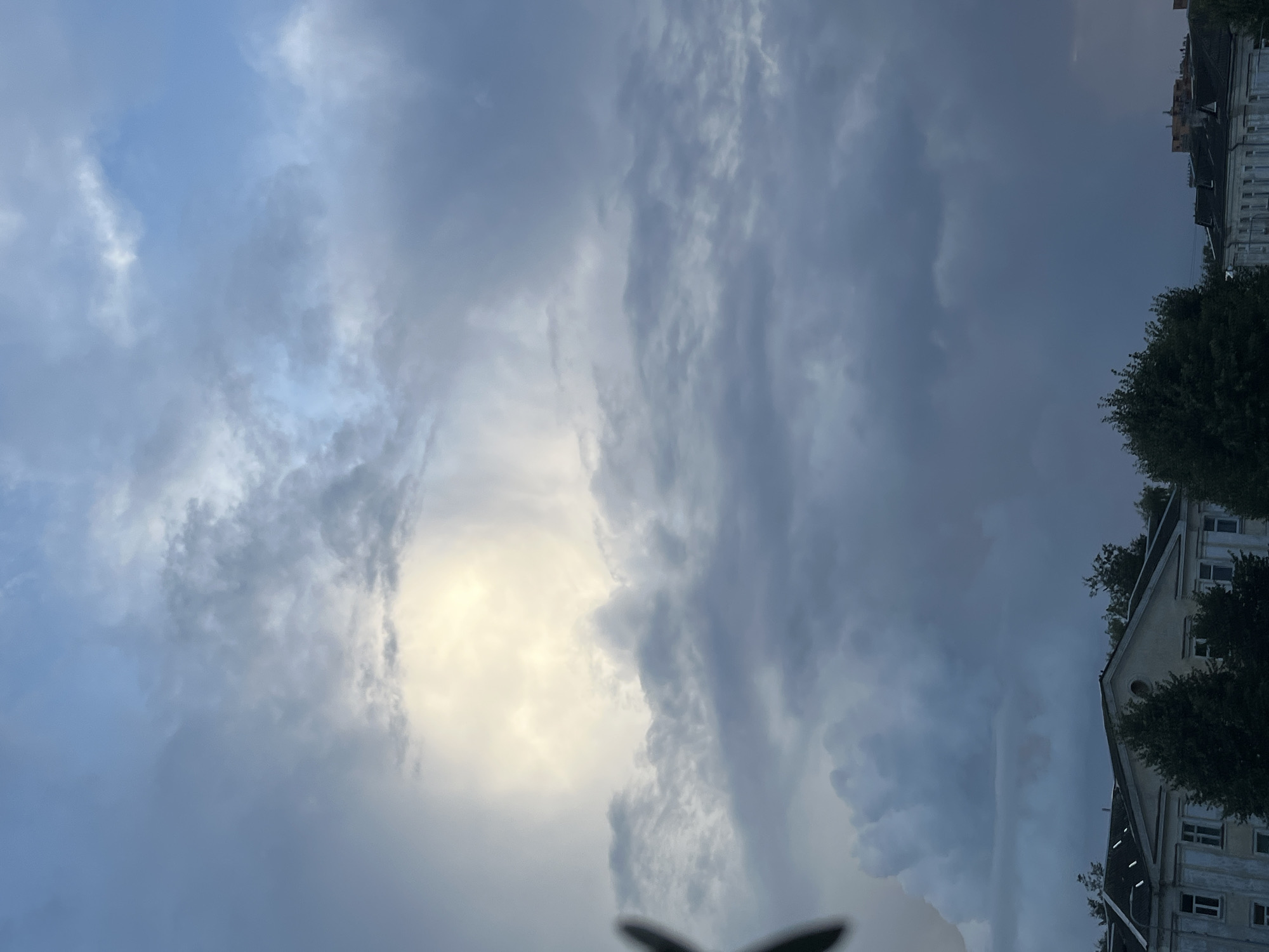 The image depicts a cloudy sky above a street in Kohtla-Järve linn, Estonia. The sky is filled with various cloud formations, ranging from dark, heavy clouds to lighter, brighter areas where sunlight is breaking through. In the lower portion of the image, the roofs of several buildings are visible, along with trees and some streetlights. The architecture suggests a mix of older and possibly more modern structures. The overall atmosphere is somewhat overcast, with the bright patch of sky indicating the presence of the sun behind the clouds. There are no people or vehicles clearly visible in the frame. The foreground on the left shows a dark, blurry object that could be foliage or an architectural element.